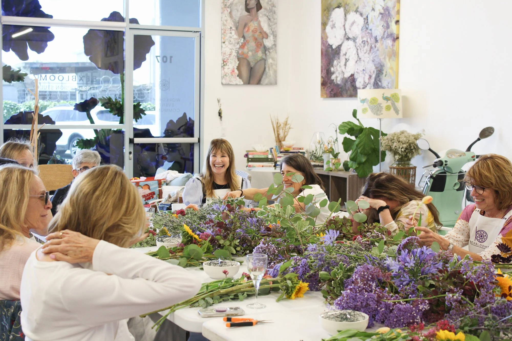 A group of women gathered around a table filled with colorful flowers, engaging in flower arrangement activity, with happy expressions, in an art-filled room.