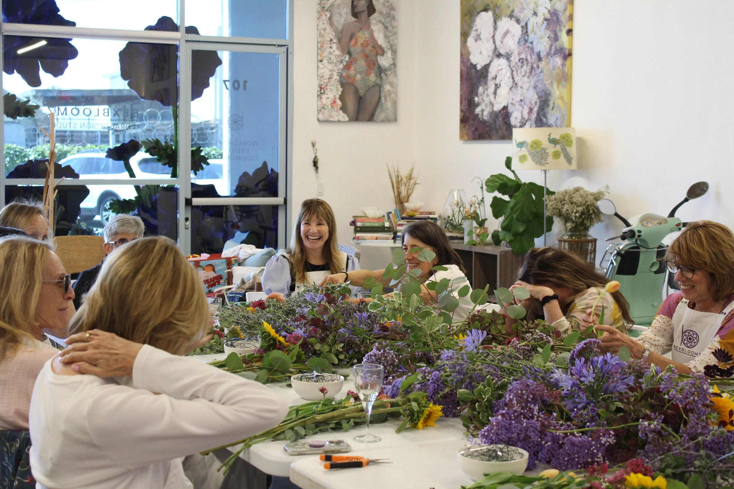 Group of people sitting around a large table decorated with an array of flowers, some smiling and engaging in a flower arrangement activity inside a room with artwork on the walls and a large window at the back.