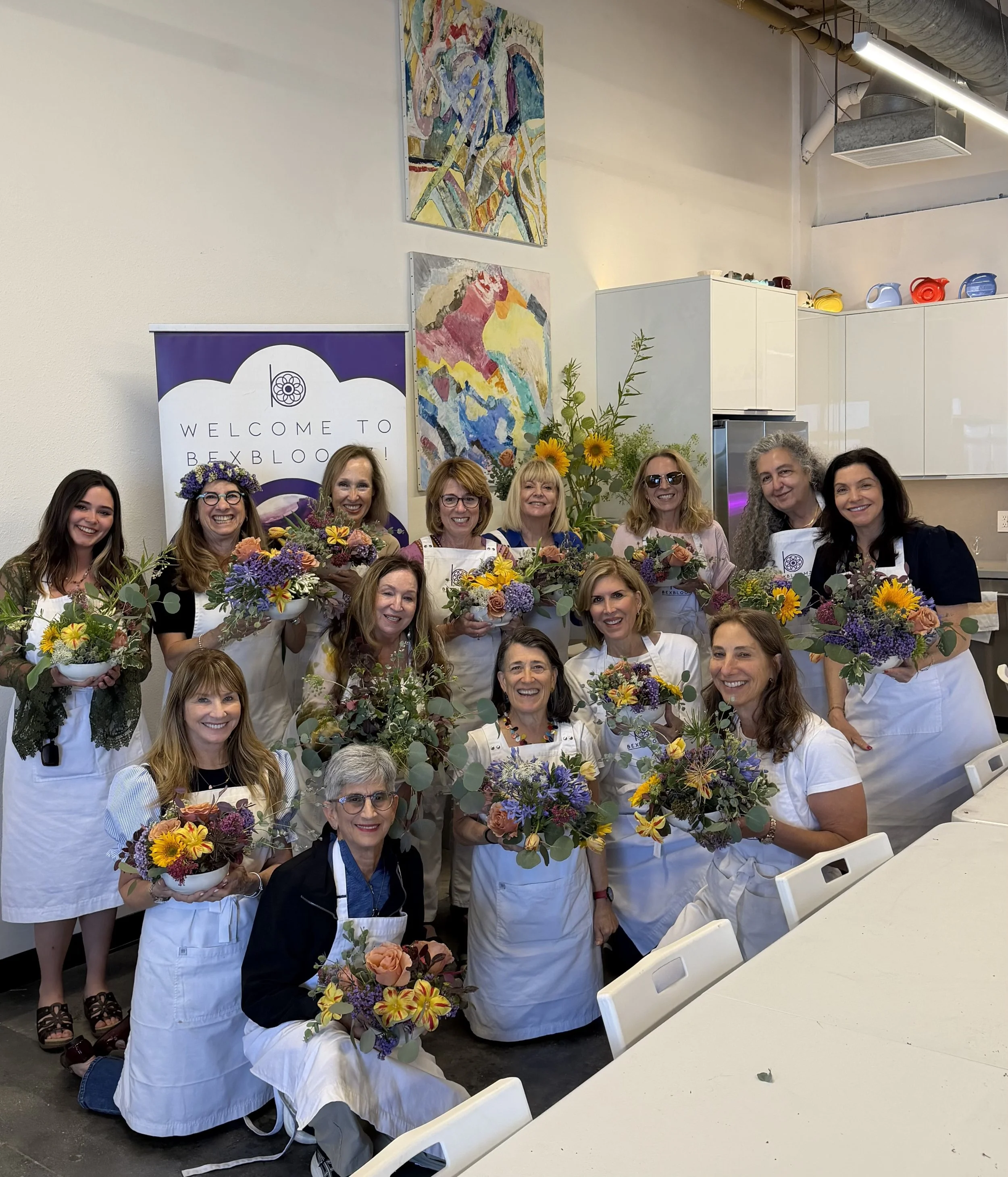 Group of women smiling and holding bouquets of colorful flowers in a room with white walls, colorful abstract paintings, and a long white table with white chairs, some women wearing white aprons, indoors at a floral arrangement event.