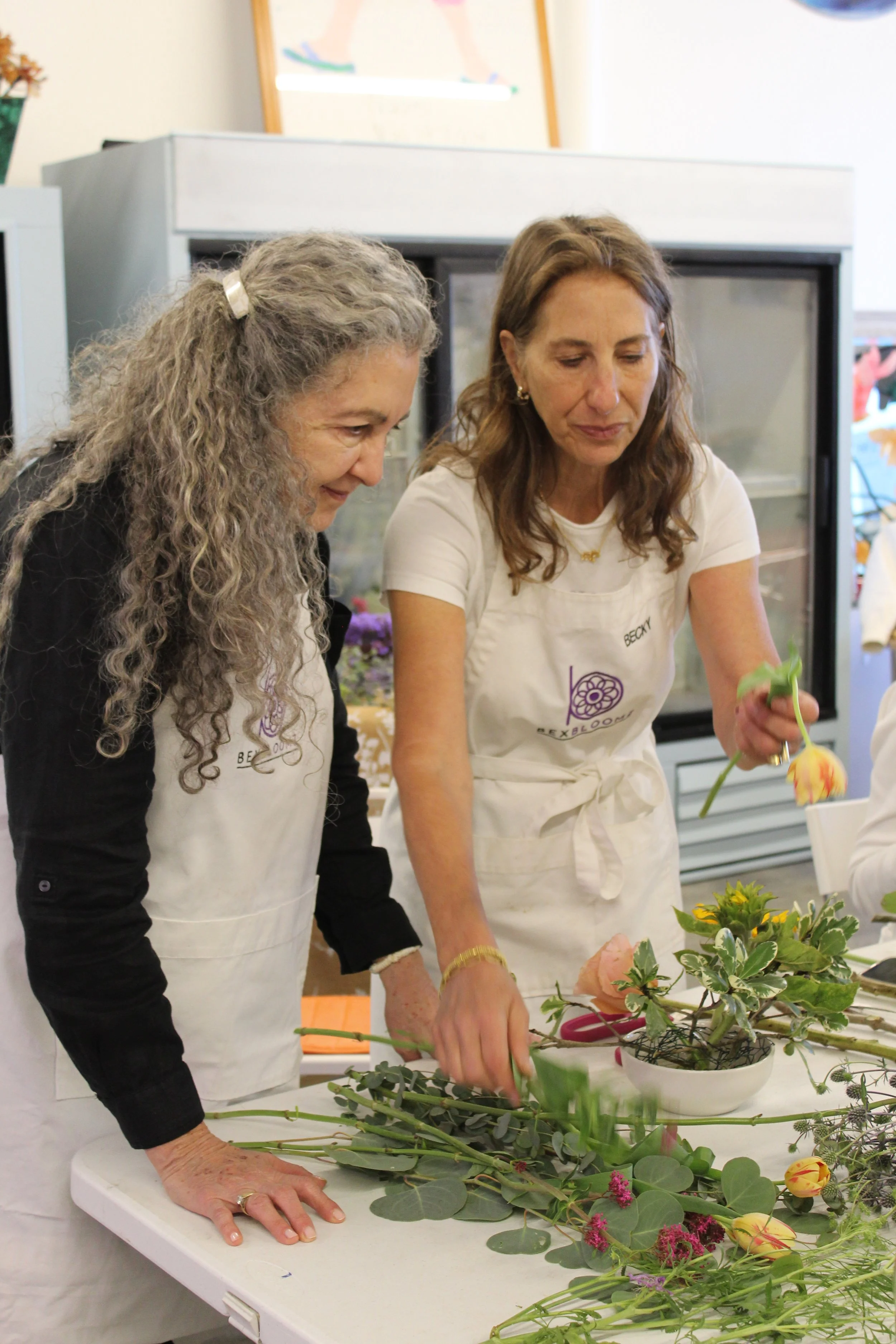 Two women wearing white aprons arranging flowers on a table. One has long curly gray hair and the other has shoulder-length brown hair. There are various flowers and greenery on the table, and a refrigerator in the background.