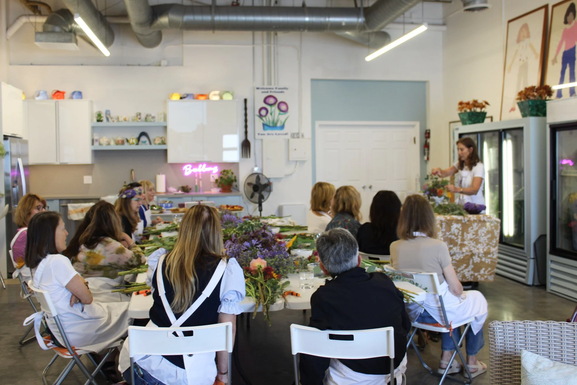 A group of people attending a flower arrangement class in a bright indoor space. There are tables with flowers and tools, and an instructor demonstrating flower arranging. The room features white walls, cabinets, framed art, and a neon sign.