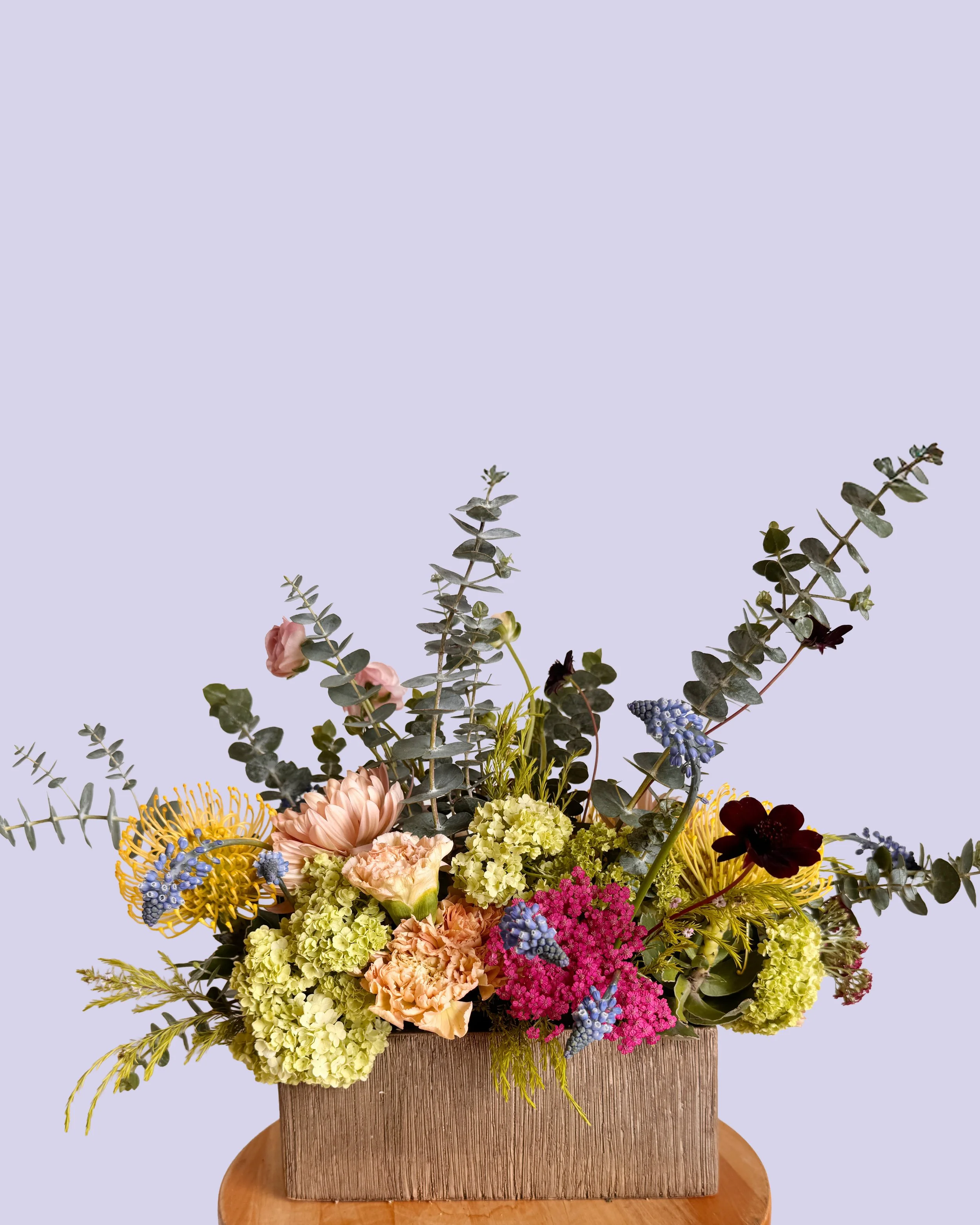 Vase filled with a variety of colorful flowers and green foliage, placed on a wooden surface against a light purple background.