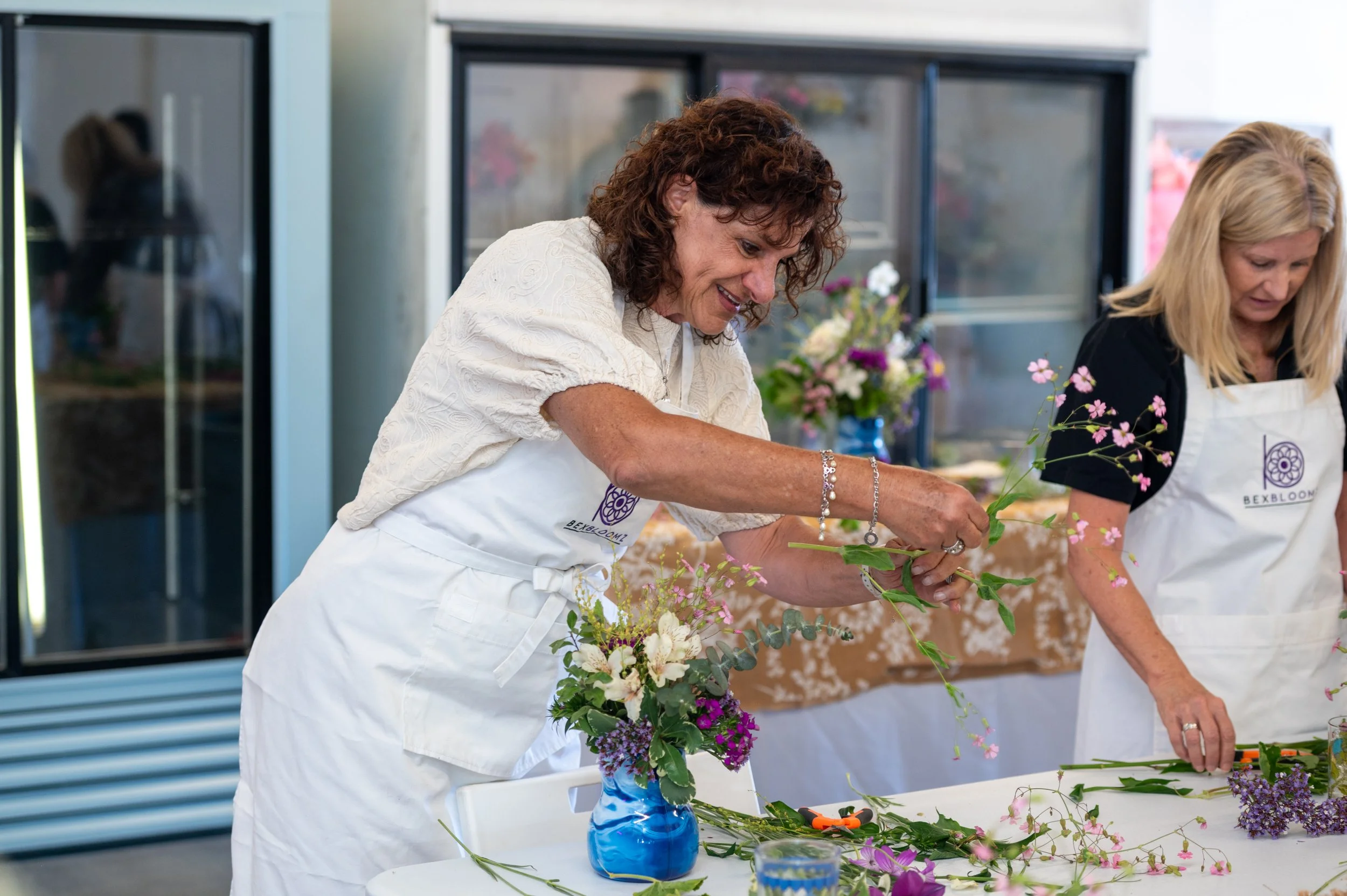 Two women arranging a floral centerpiece at a table, with flowers and a blue vase visible on the table, in a bright indoor setting.