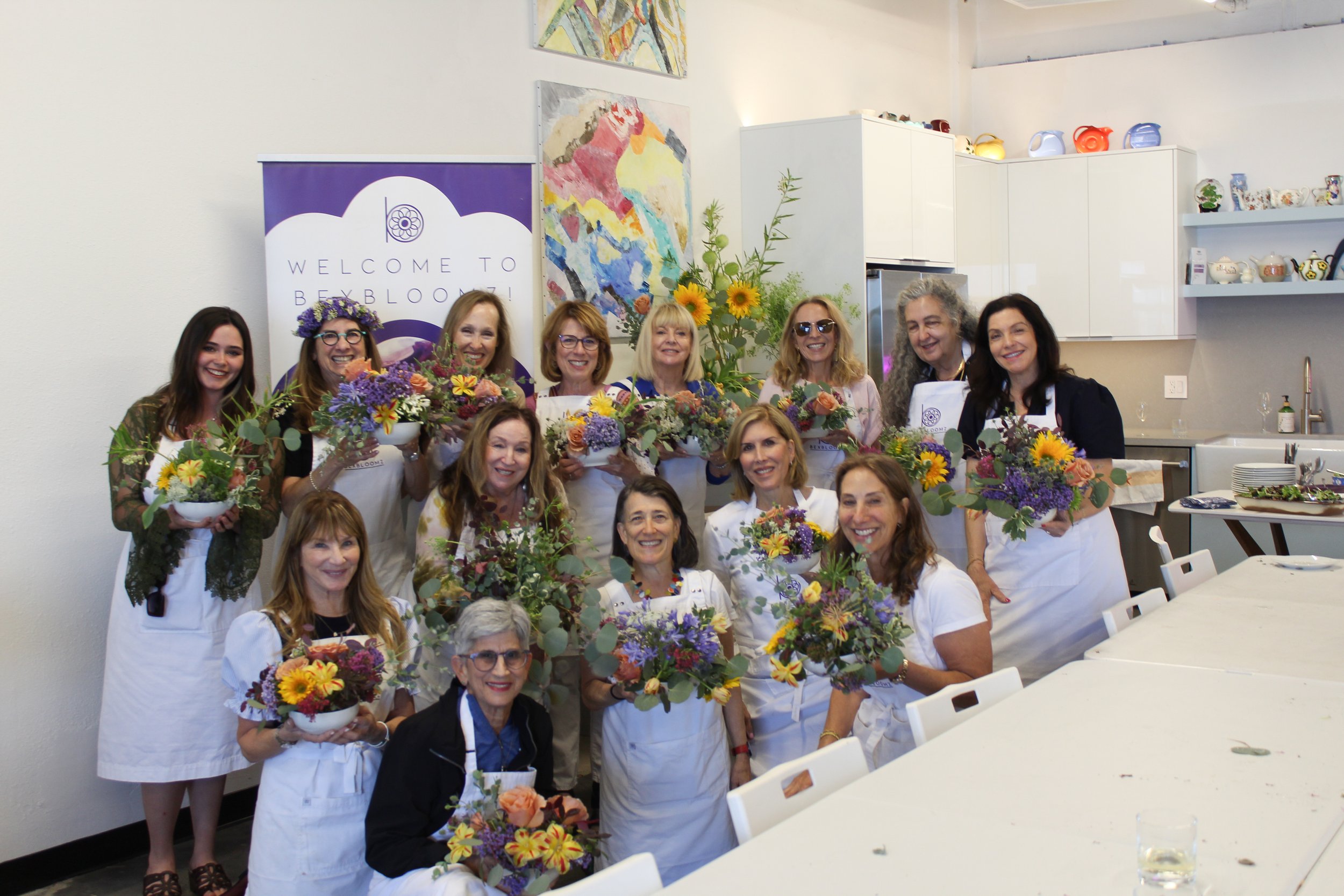 A group of women gathered in a bright room, smiling and holding bowls of colorful flowers, with a sign that says 'Welcome to Beybloom' in the background, along with abstract artwork and kitchen shelves with various teapots and decorative items.