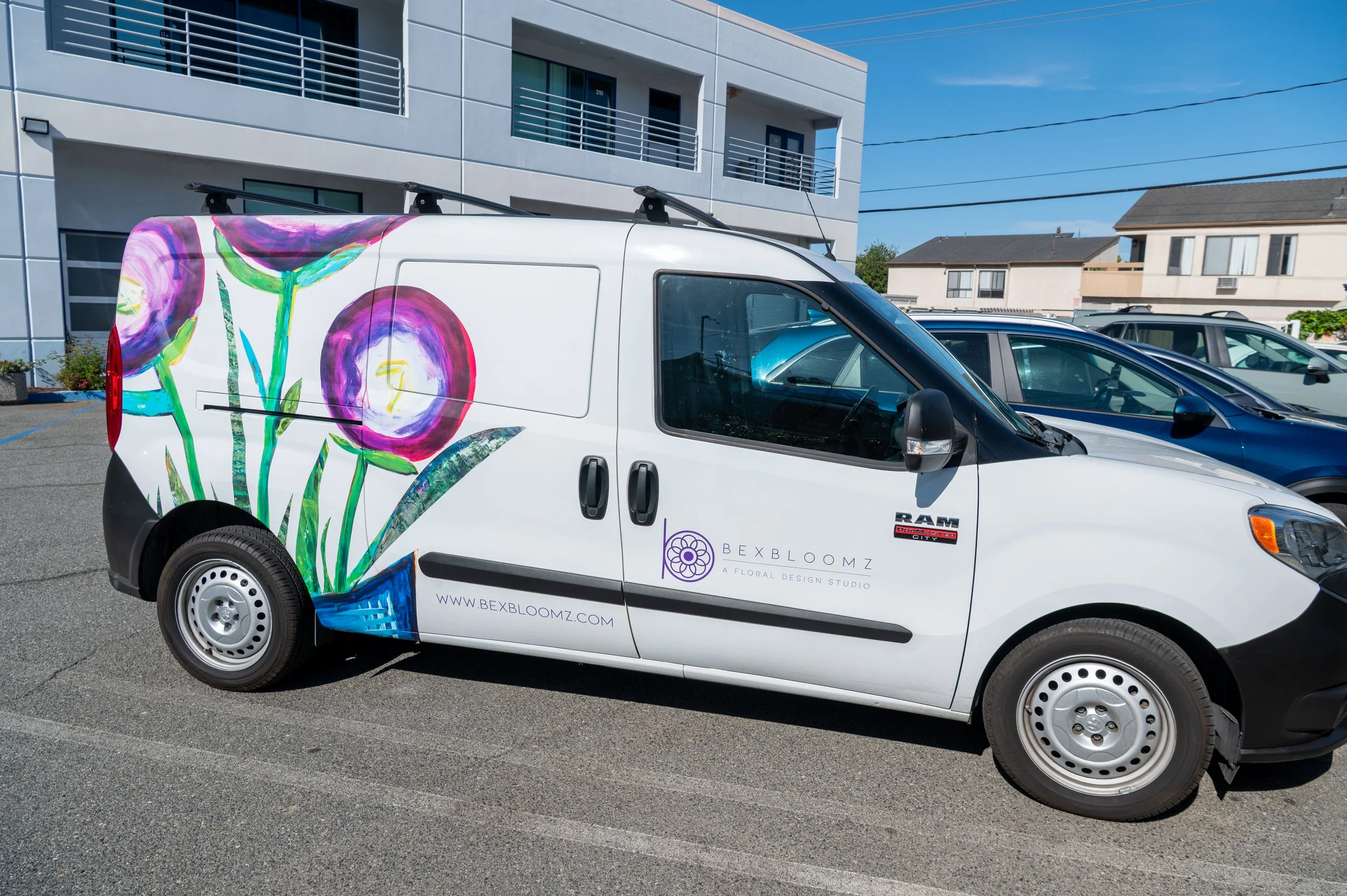 A white commercial van with colorful floral artwork on its side, parked in a parking lot near modern residential buildings on a sunny day.