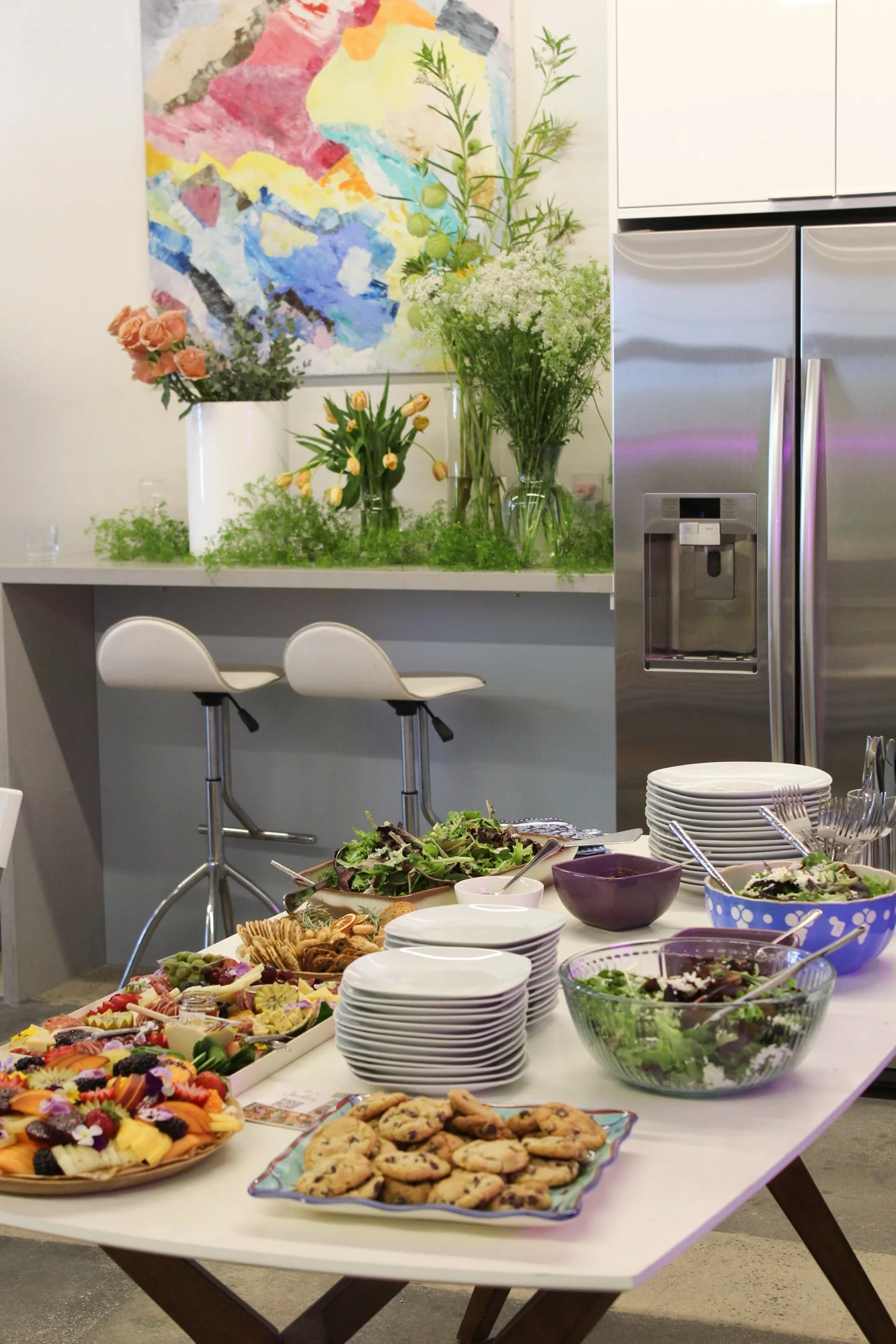 A table filled with various desserts, salads, and snacks at a gathering, with additional floral arrangements and a kitchen in the background.