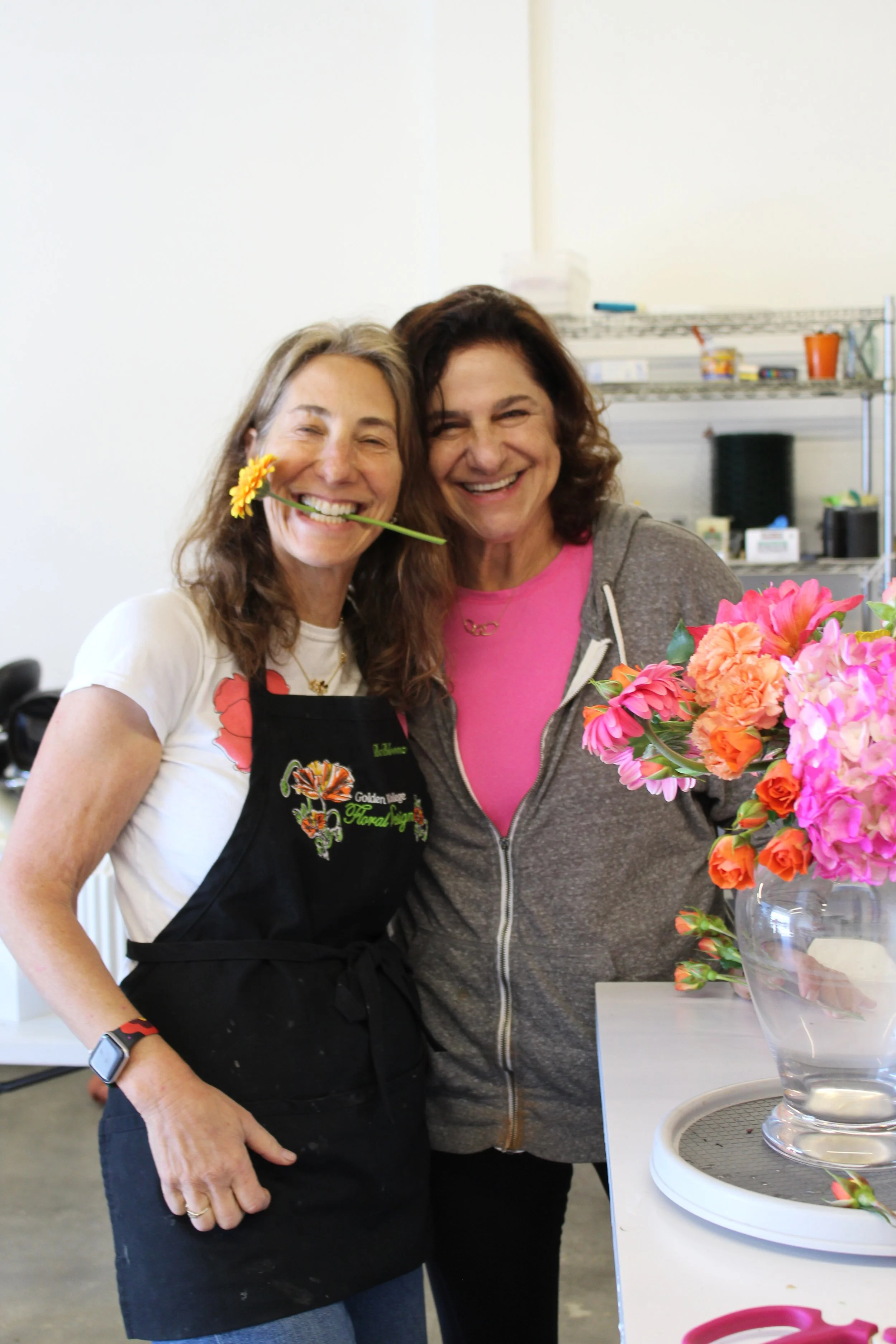 Two women smiling, one with a flower in her mouth, standing near a table with a large bouquet of pink and orange flowers in a glass vase.