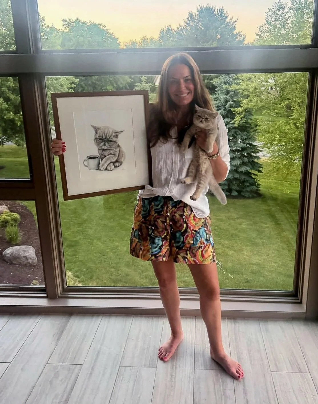 A woman holding a framed cat illustration and a real cat in front of large windows with trees outside