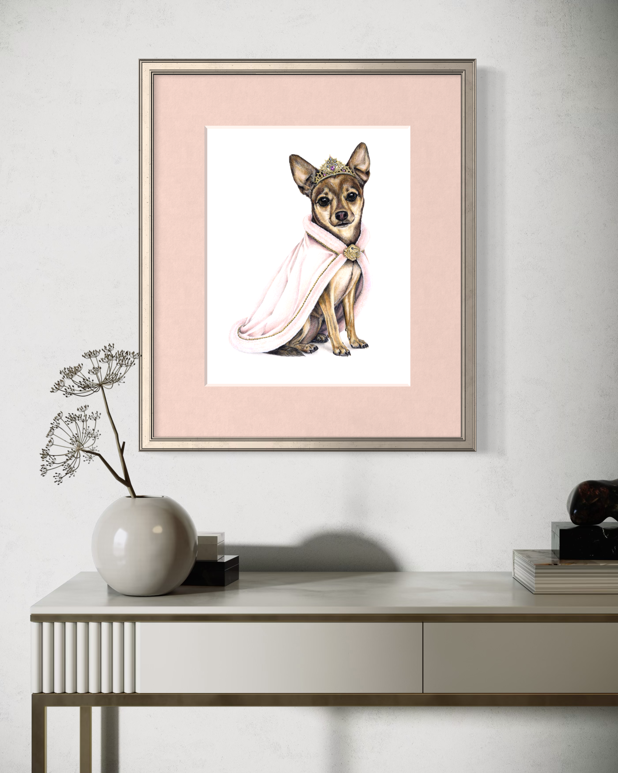 A minimalist modern sideboard with a large, round white vase containing a single dried flower on the left, and a small black cube and a glass decor item on the right. Above, there's a framed artwork of a small dog wearing a princess crown and pink cape, against a light pink background.