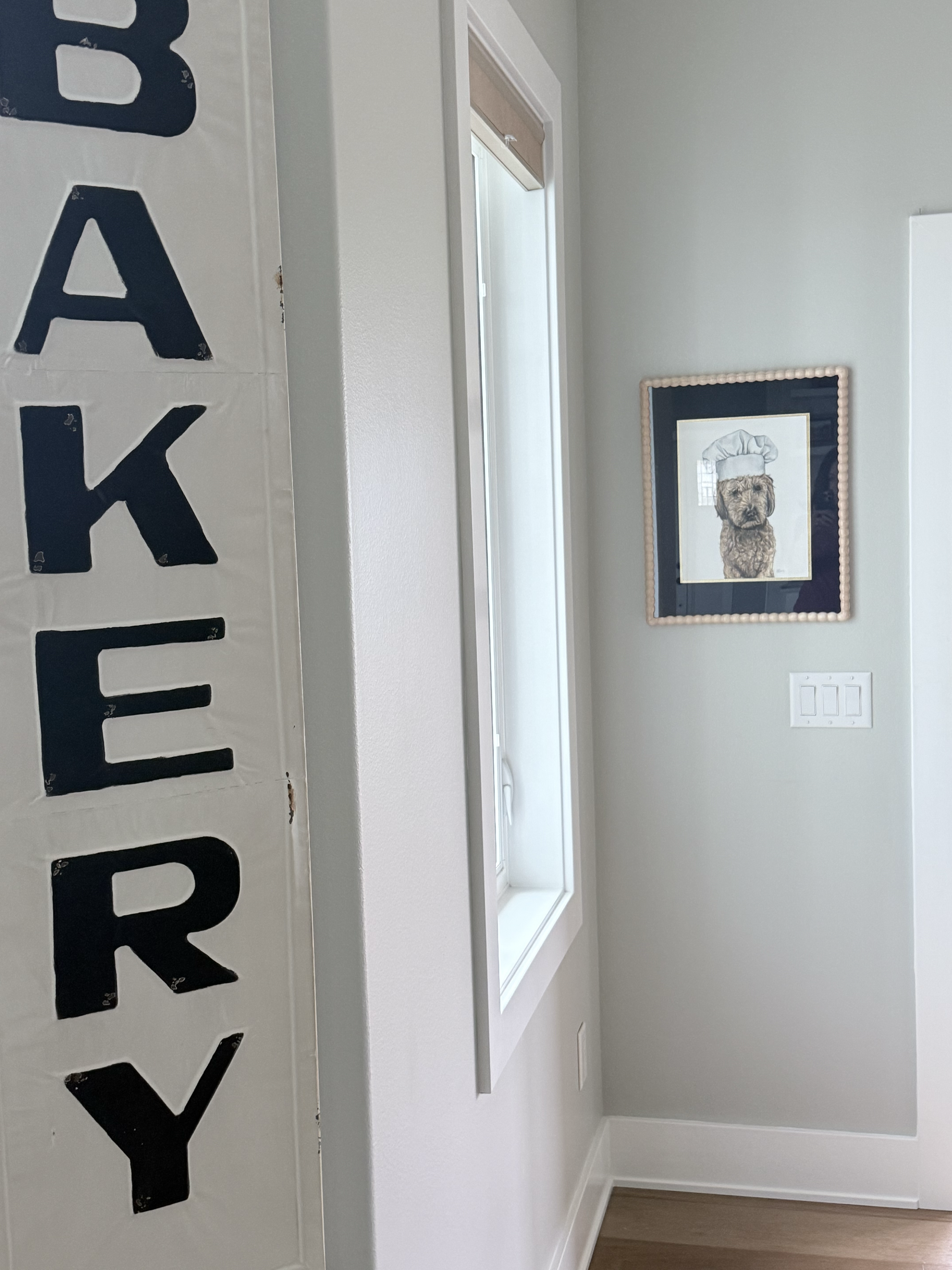 A white countertop with a partially visible black and white sign that has large black letters, and a framed picture of a dog dressed as a chef on the wall in a room with light gray walls and a window.