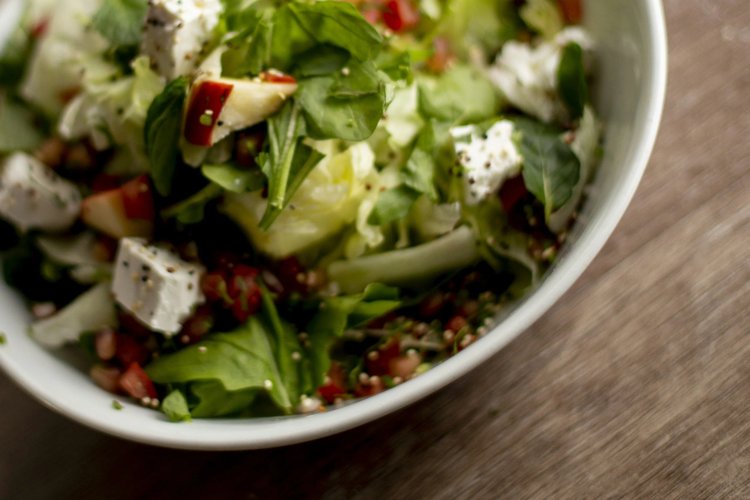 Close-up of a fresh vegetable salad in a white bowl, containing leafy greens, red bell peppers, feta cheese, and seeds, on a wooden surface.