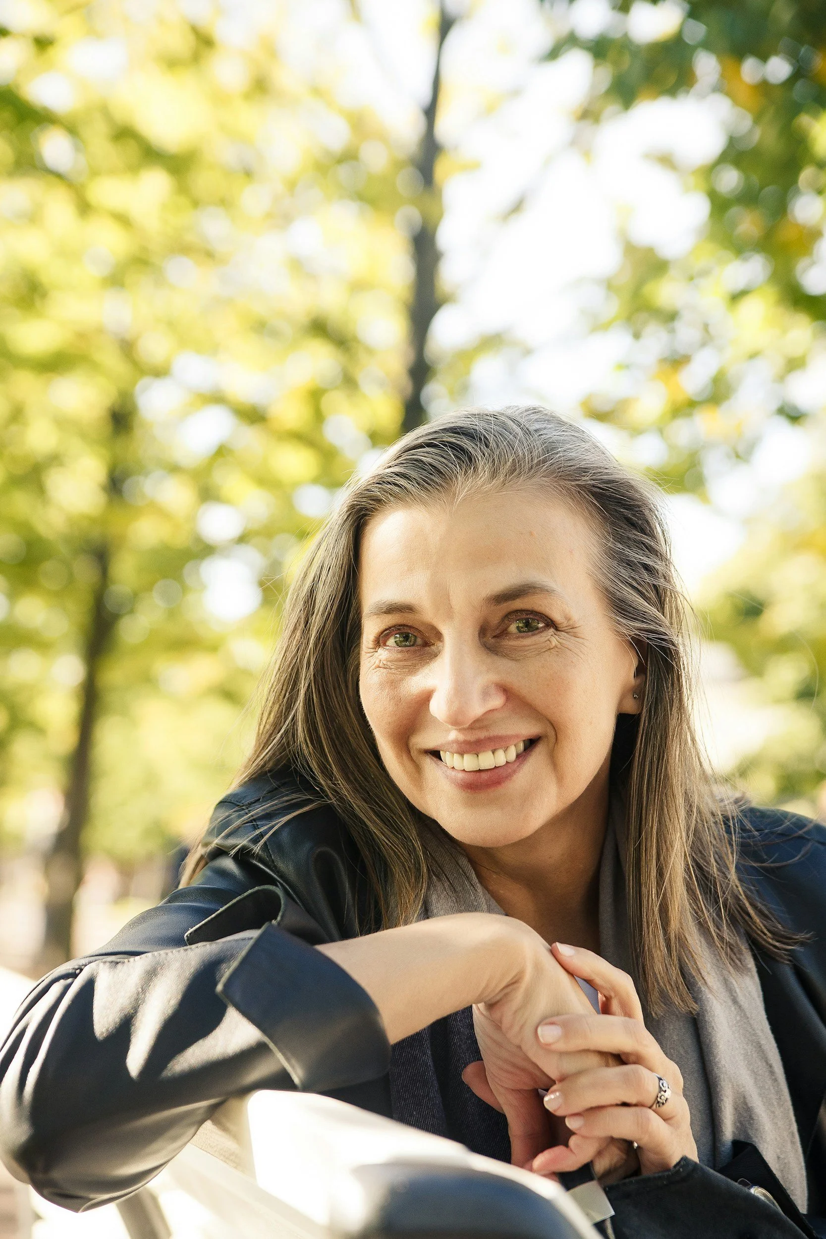 A woman smiling outdoors with yellow and green blurred leaves in the background, wearing a dark jacket, gray shirt, and jewelry, leaning forward with her arms resting on something in front of her.