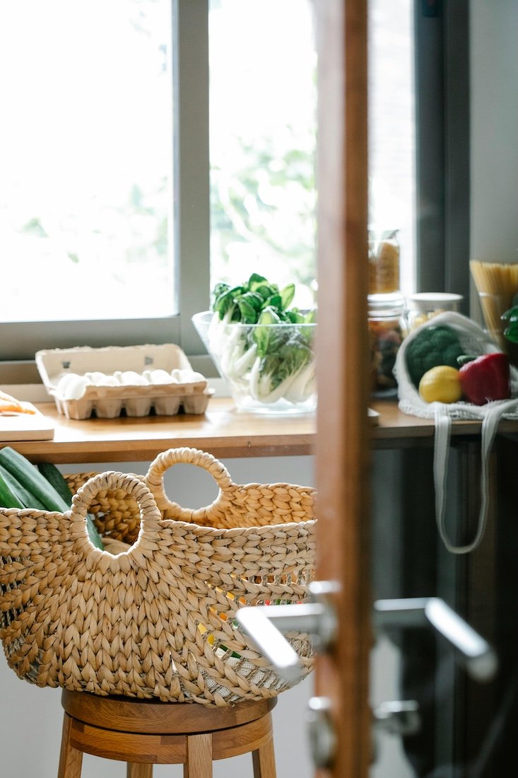 Fast forward nutrition -  A woven basket with green zucchinis sitting on a wooden stool in front of a kitchen window. In the background, there are fresh vegetables, eggs, and leafy greens on a countertop.