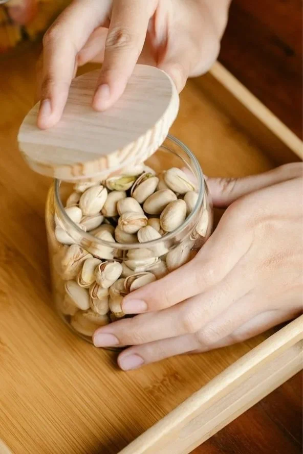 Whole food approach to healthy eatingPerson opening a jar of pistachio nuts with wooden lid on a wooden tray.