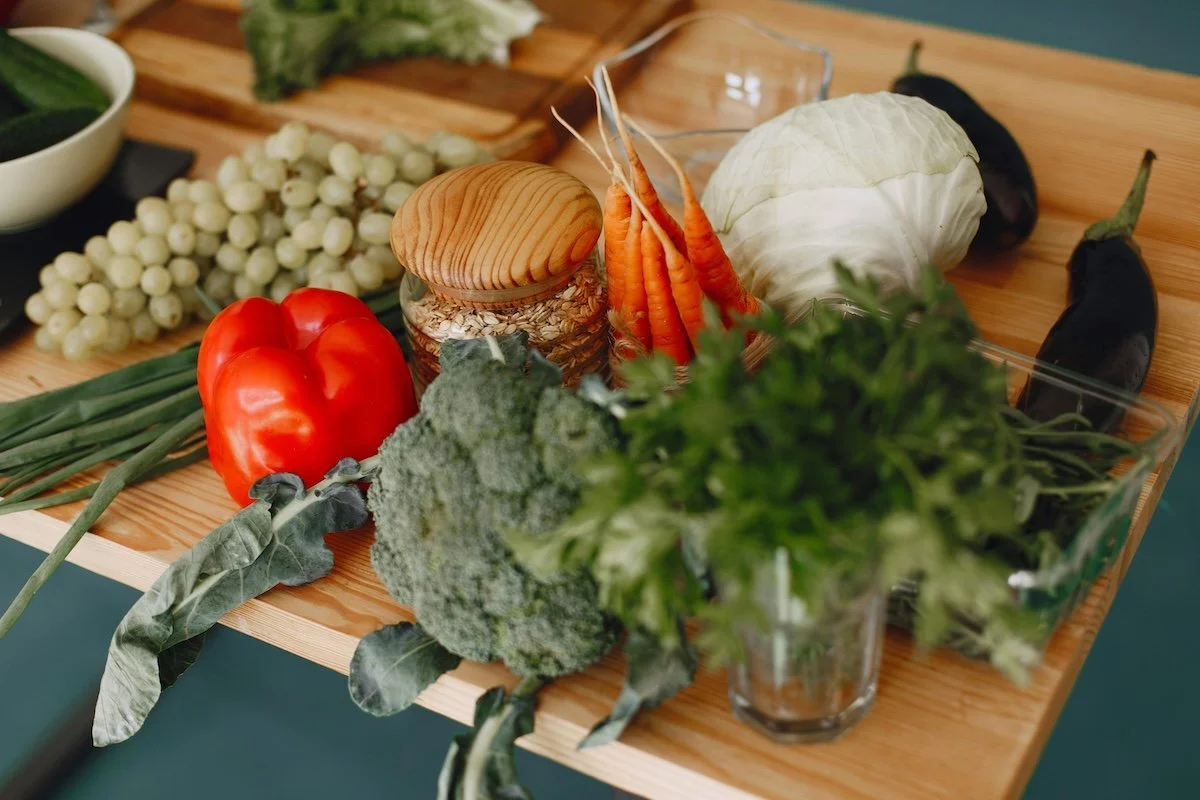 Assorted vegetables on a wooden table, including broccoli, tomato, green beans, carrots, cabbage, eggplants, and leafy greens, with a jar of oats and a bowl of green liquid in the background.