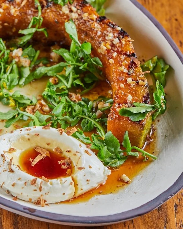 Roasted pumpkin slice topped with herbs and cheese, served with a soft-boiled egg drizzled with soy sauce, all on a bed of fresh greens