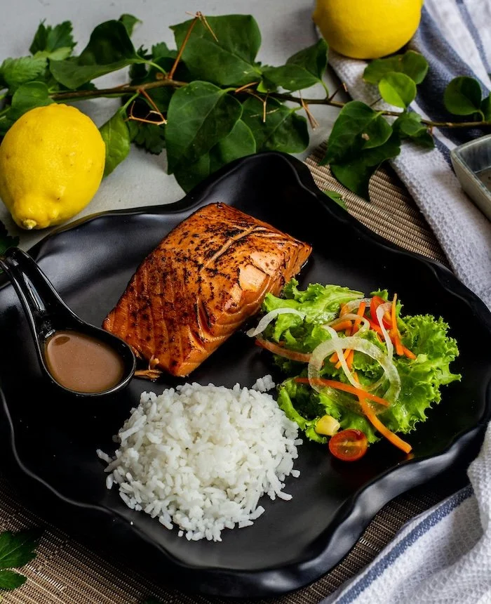 Grilled salmon fillet with a side of white rice, fresh salad with lettuce, cherry tomatoes, shredded carrots, and onion, on a black plate, decorated with green leaves and lemons in the background.