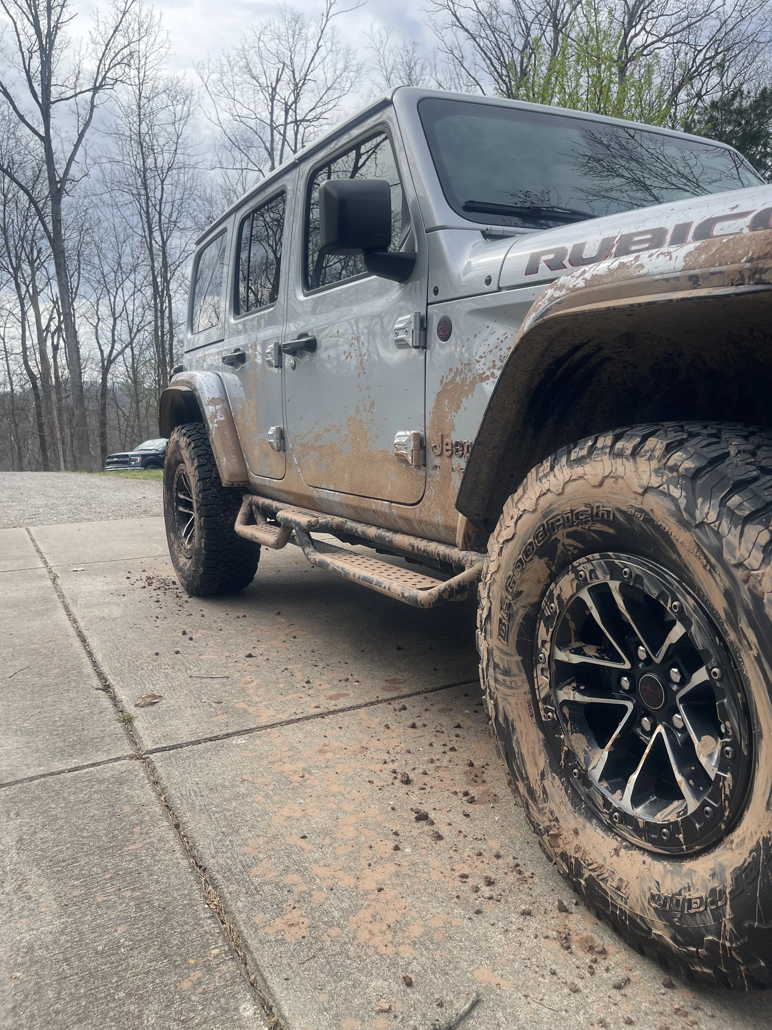 A silver Jeep Rubicon parked on a concrete driveway with mud splattered on its body and tires, surrounded by trees.