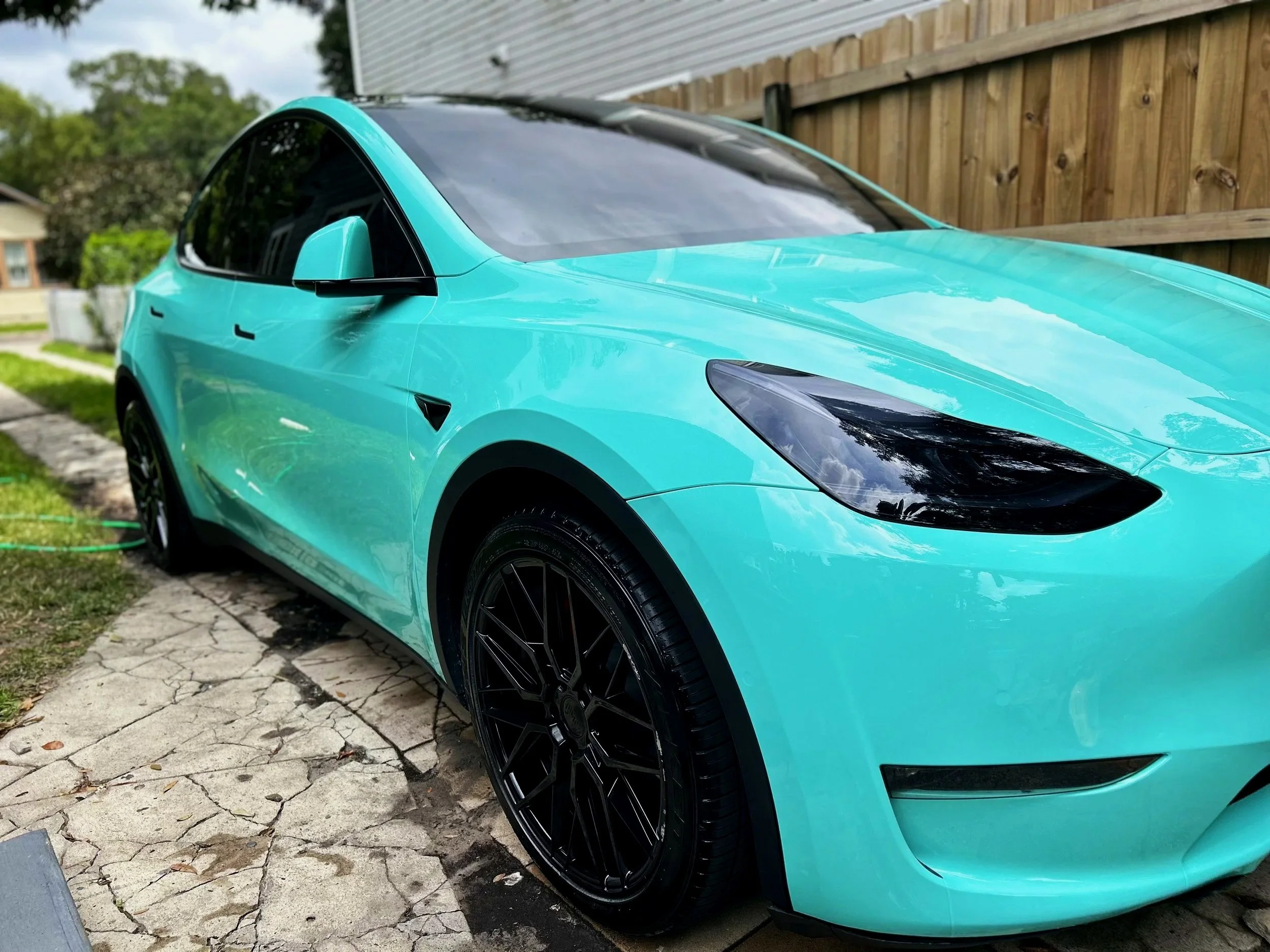 A turquoise Tesla Model 3 parked outside next to a wooden fence on a stone driveway.