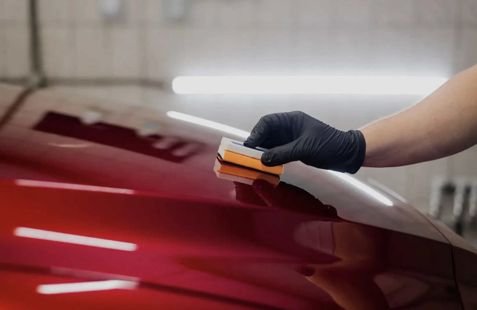 A person wearing a black glove holding an orange polishing pad is polishing the red exterior of a car in a garage or workshop.