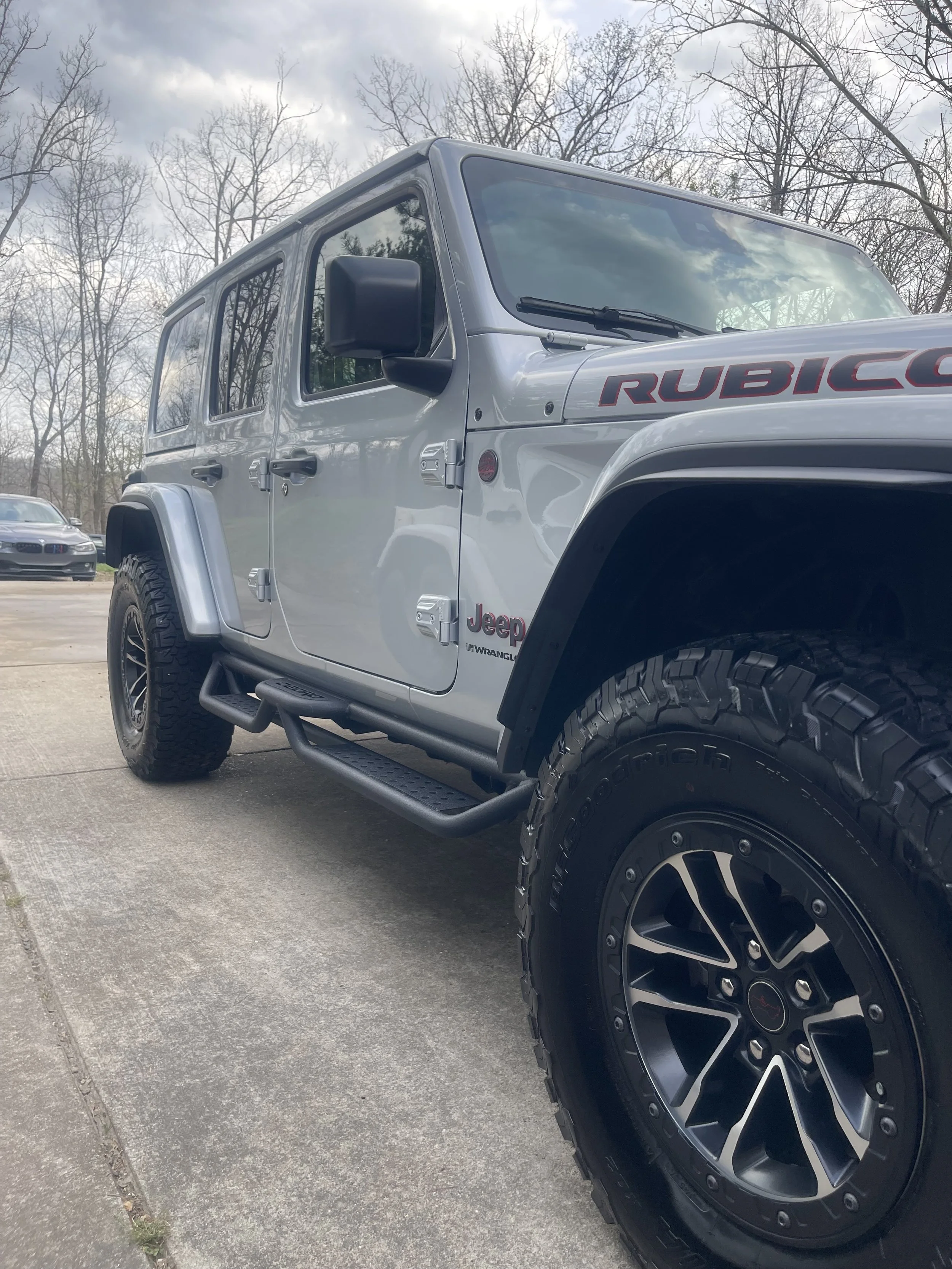 A silver Jeep Wrangler Rubicon parked on a concrete surface with trees in the background.