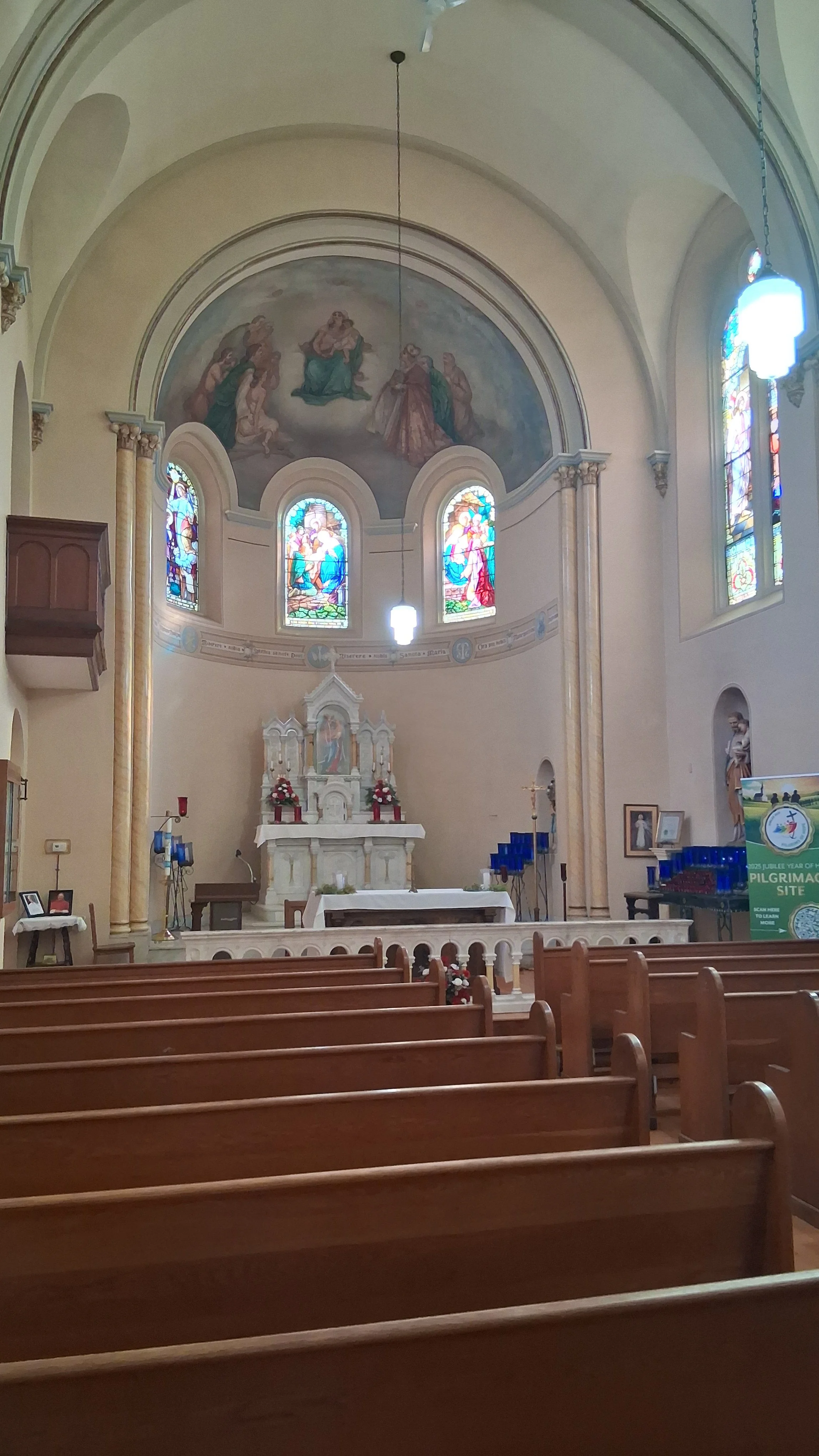 Interior of a church with wooden pews, stained glass windows, an altar with flowers, and religious artwork on the walls and ceiling. Shrine of our Lady of Sorrows, Rhineland, MO.
