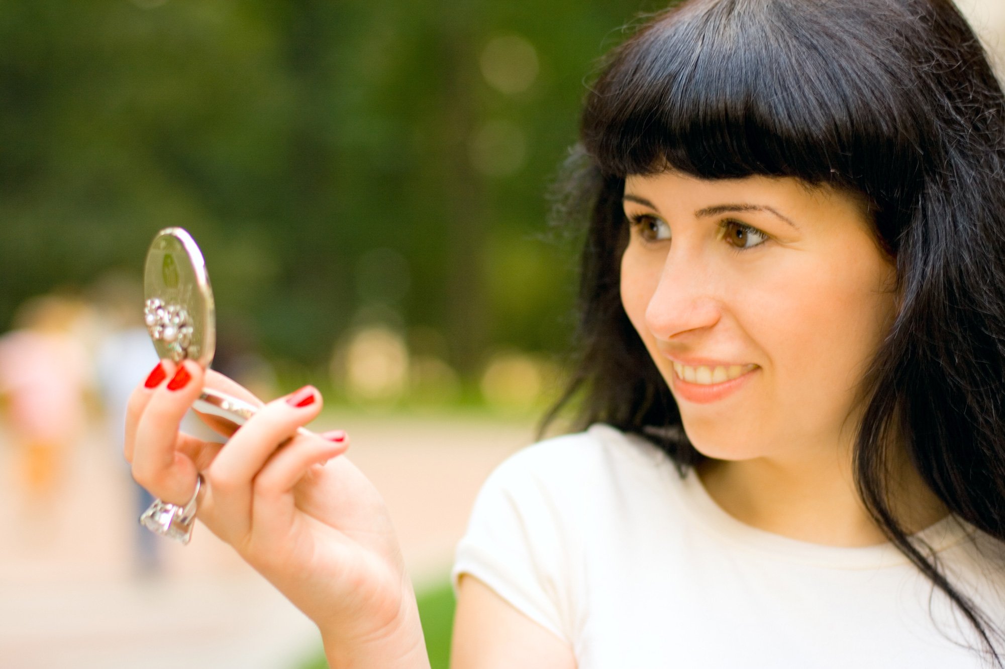 A young woman with dark hair and light skin looks in a mirror she is holding with her right hand, smiling in an outdoor setting with blurred greenery in the background.