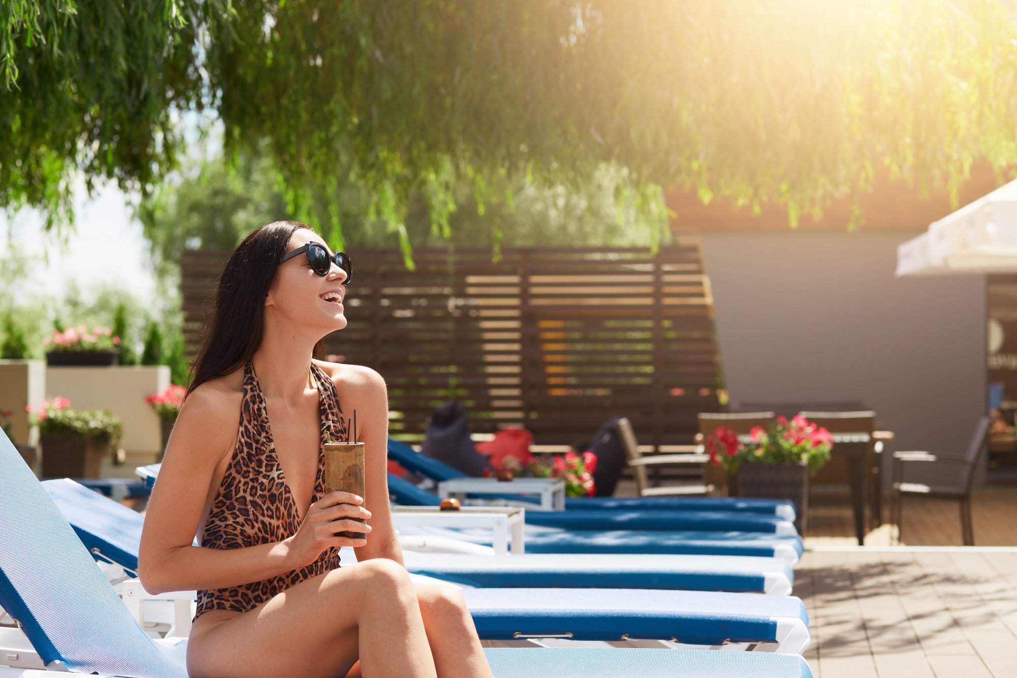 A woman with long dark hair, wearing a leopard print swimsuit and sunglasses, sitting by a pool with a drink in hand.