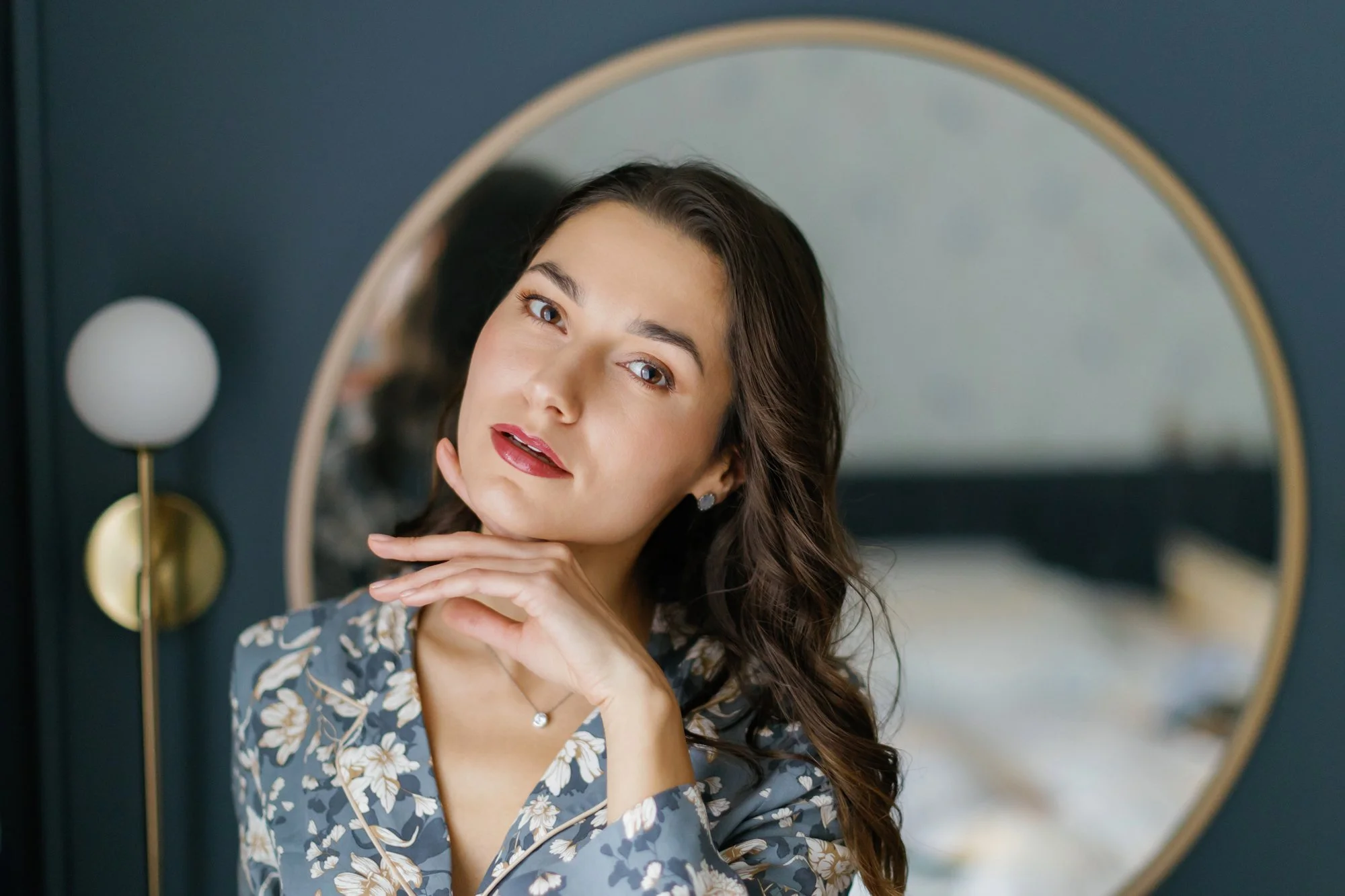 A woman with long brown hair posing in front of a mirror, touching her chin with her hand, wearing a floral blouse.