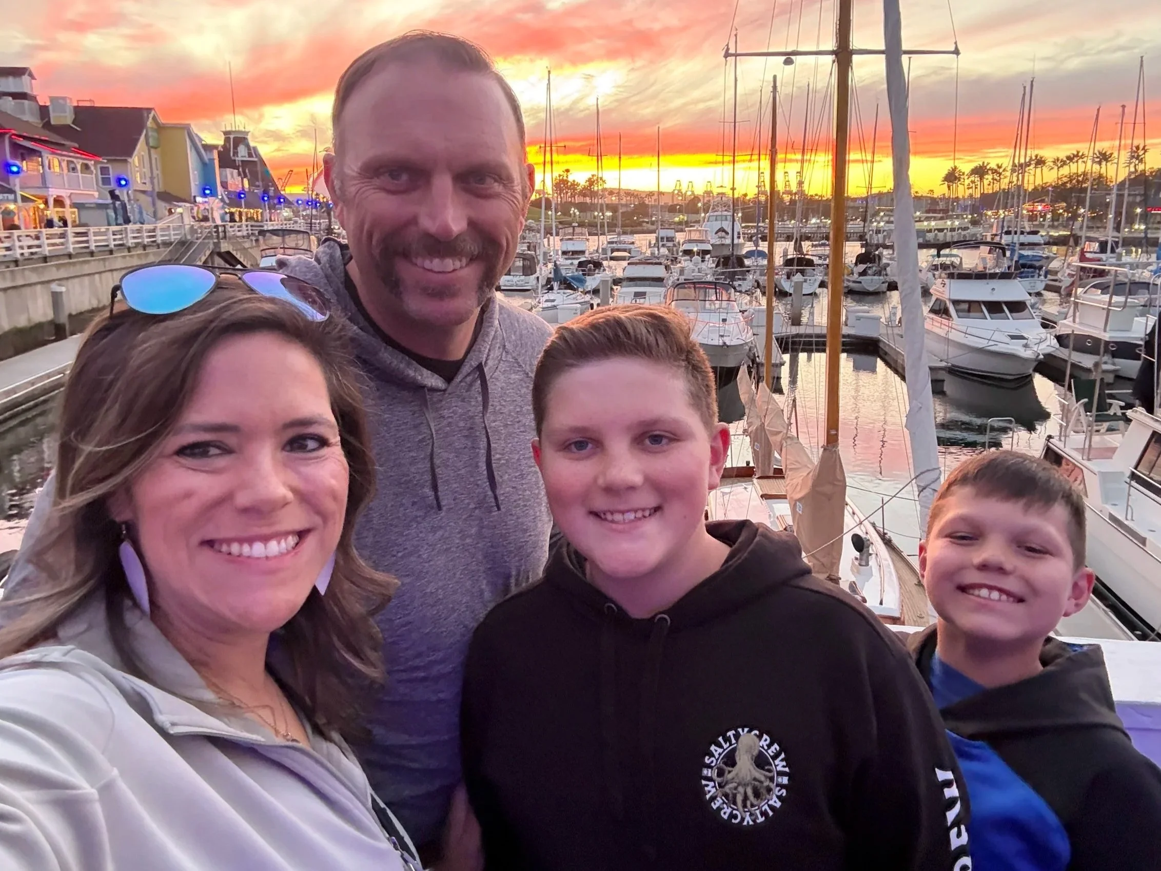 A family of four smiling at the camera at a marina during sunset, with boats and colorful sky in the background.