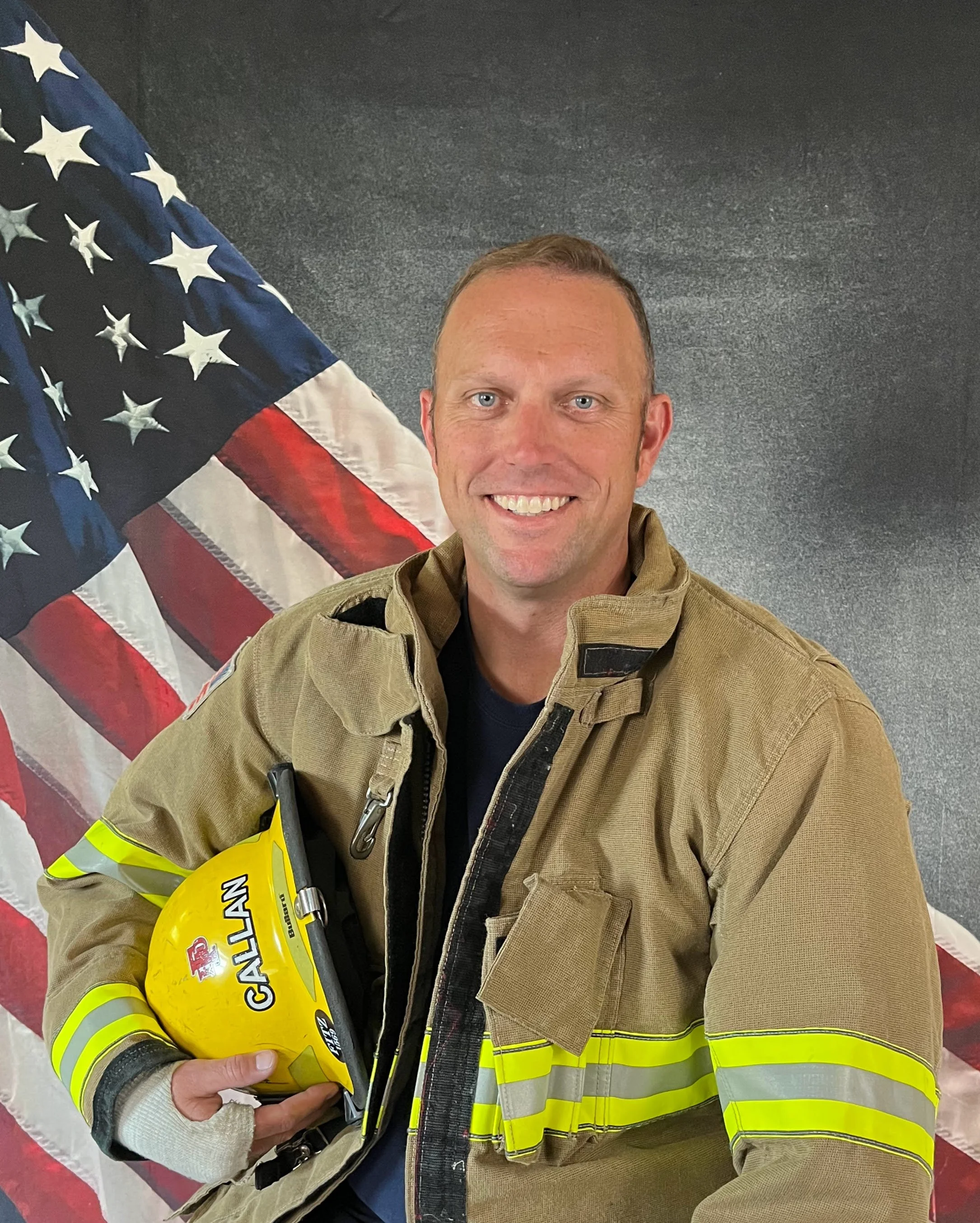 A smiling firefighter holding a yellow helmet in front of an American flag.