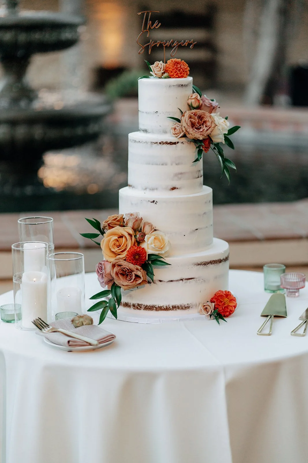 A four-tier naked wedding cake decorated with pink, peach, and orange flowers, with a "The Suppliers" cake topper, on a round table with candles and tableware.