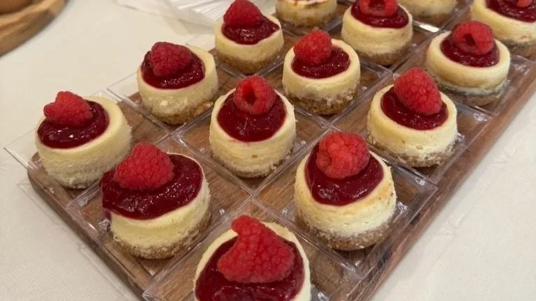 Small round cheesecakes topped with raspberry and raspberry sauce, arranged on a clear plastic tray.