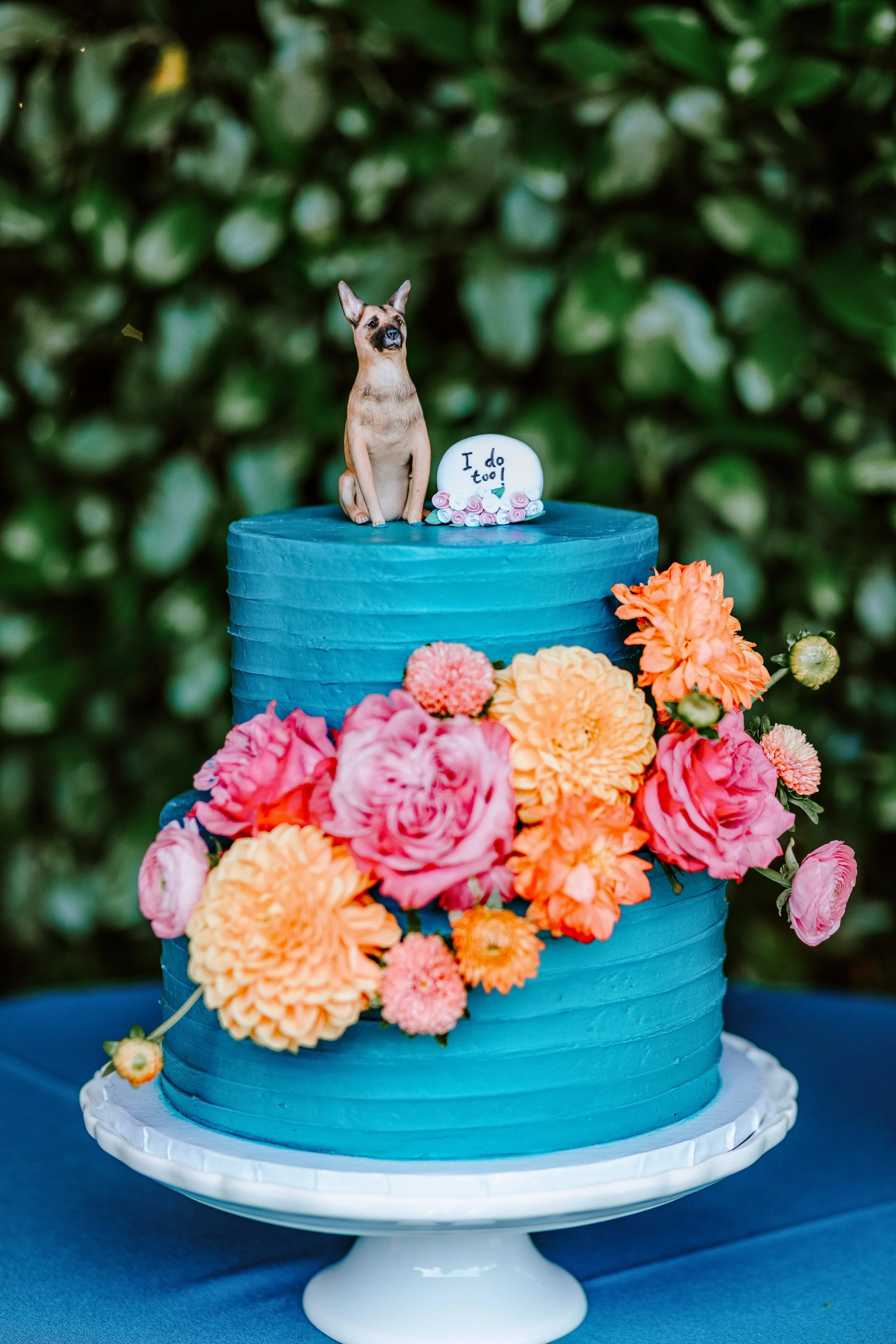 A two-tiered blue cake decorated with pink and orange flowers around the sides, topped with a dog figurine and a white fondant plaque that reads "I do too!"