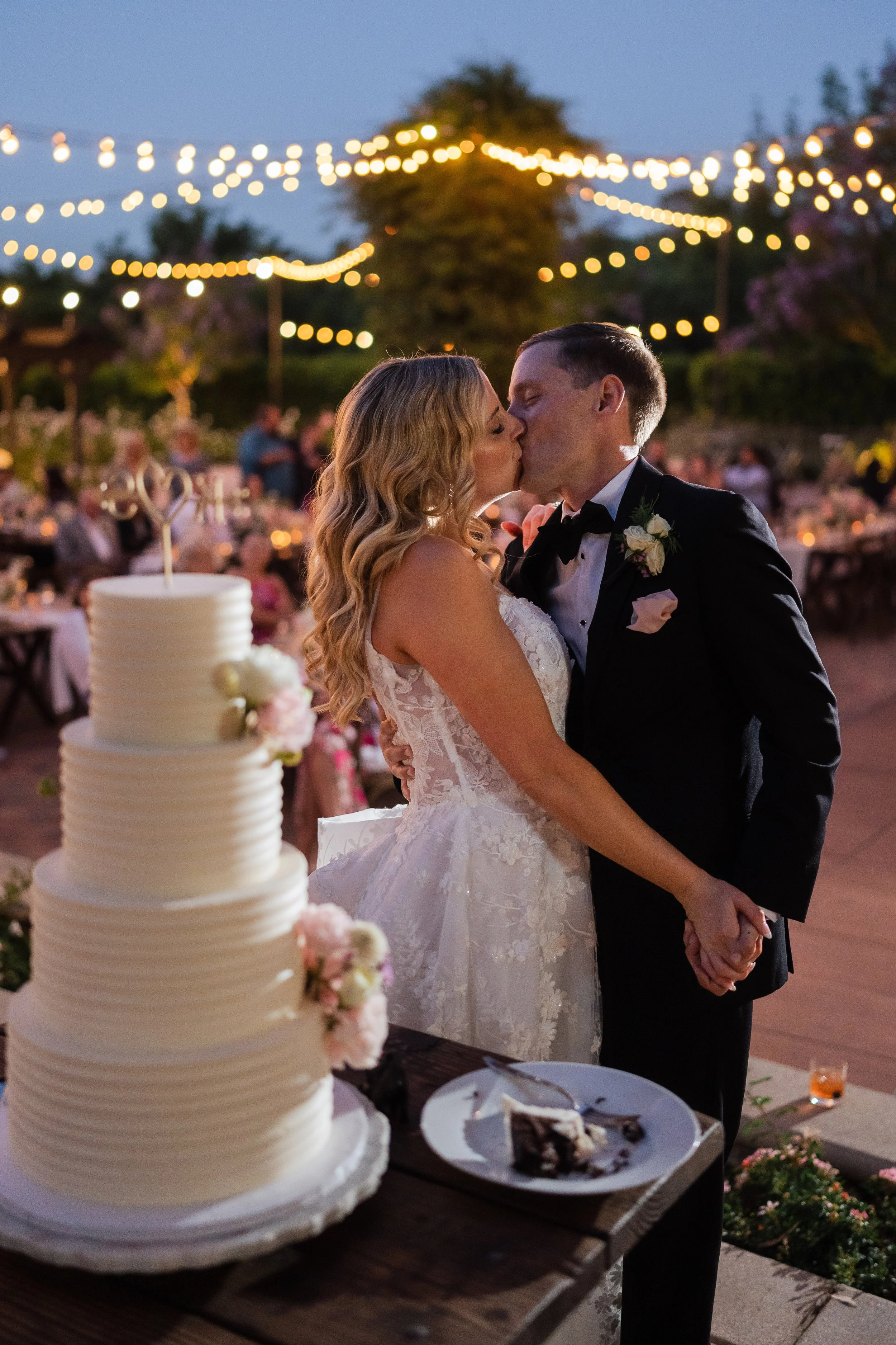 A bride and groom kissing during their wedding reception at night, decorated with string lights, with a wedding cake and dessert table in the foreground.