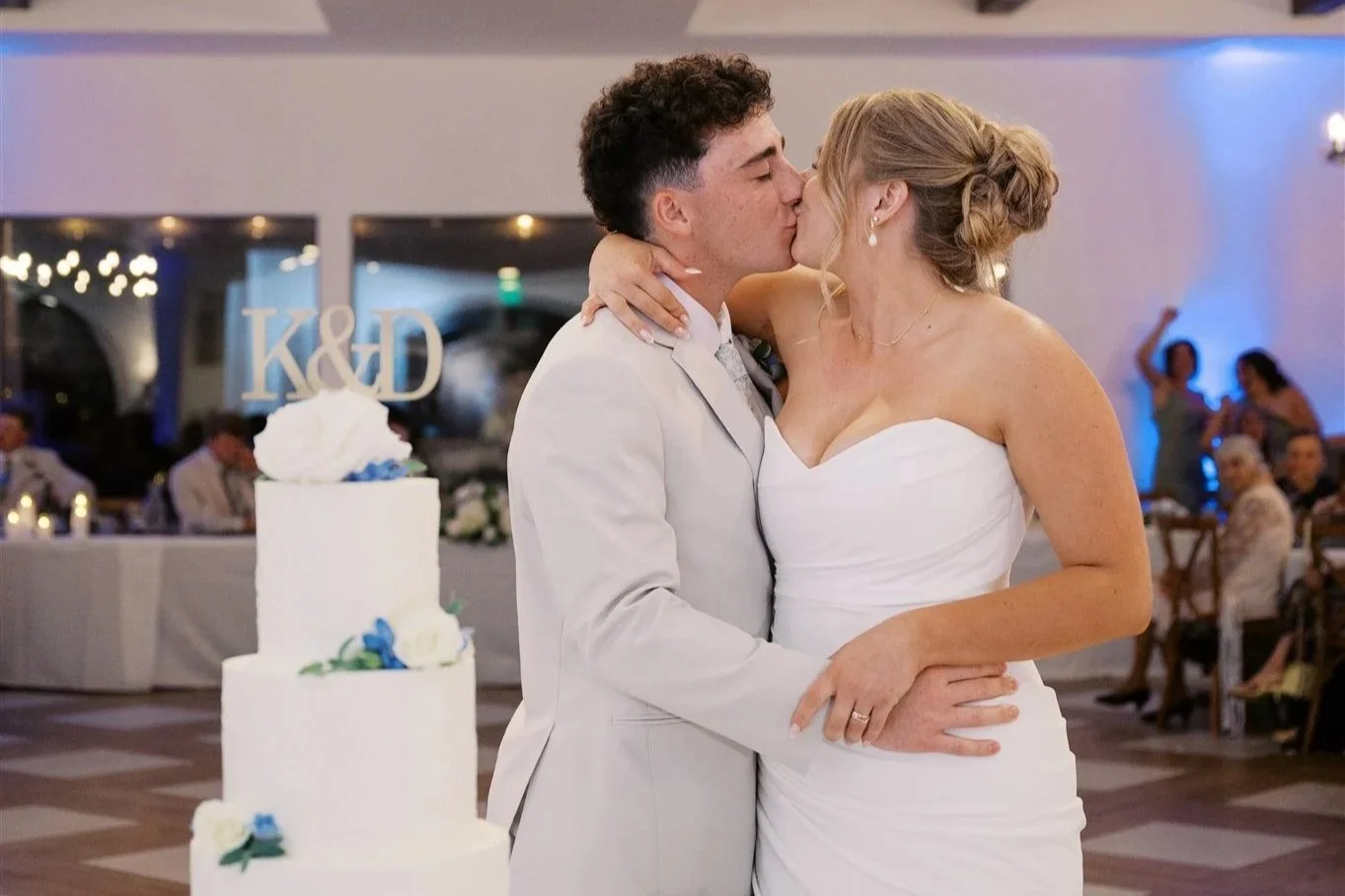 A bride and groom share a kiss during their wedding reception, with a wedding cake in the foreground and guests seated at tables in the background.