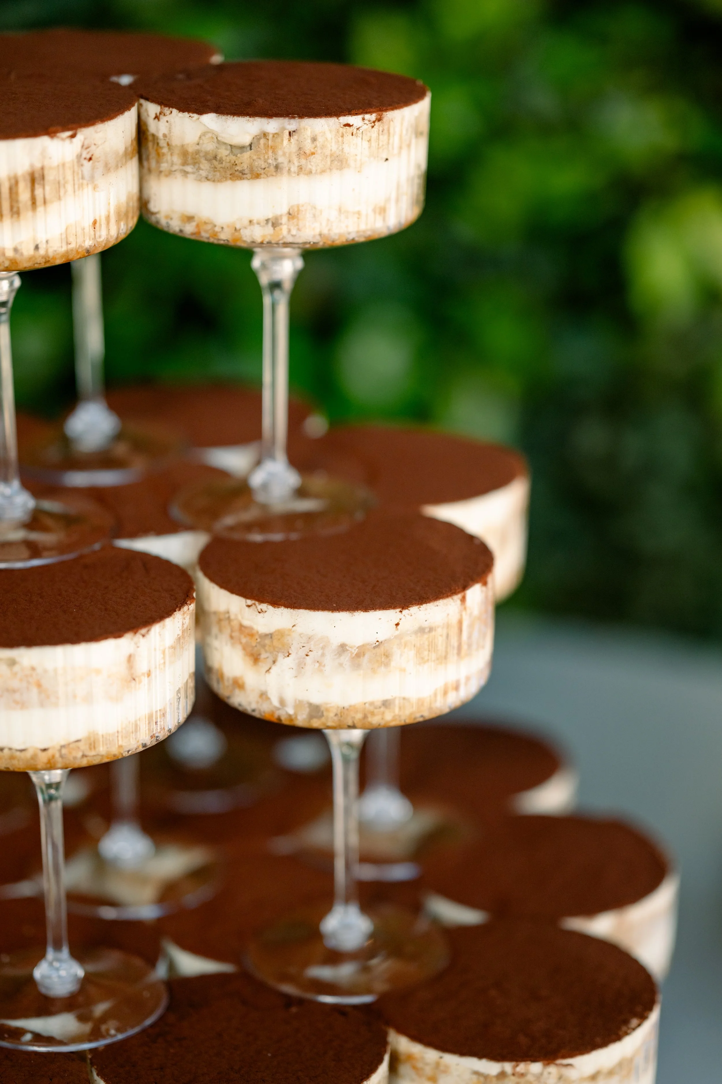Close-up of a multi-layered dessert with a chocolate top layer, served in clear glasses on a tiered stand, with a blurred green background.