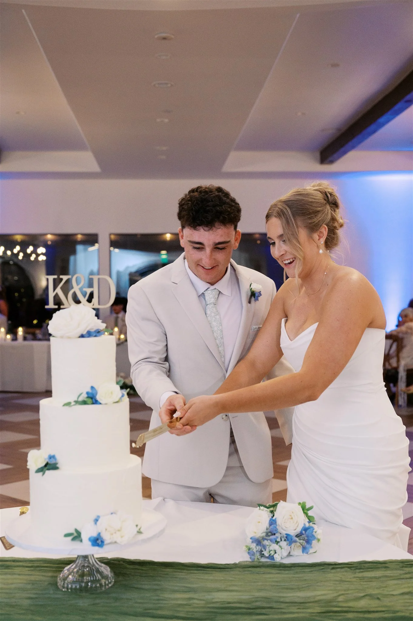 A wedding couple, a groom and bride, cutting their wedding cake together in a decorated venue.