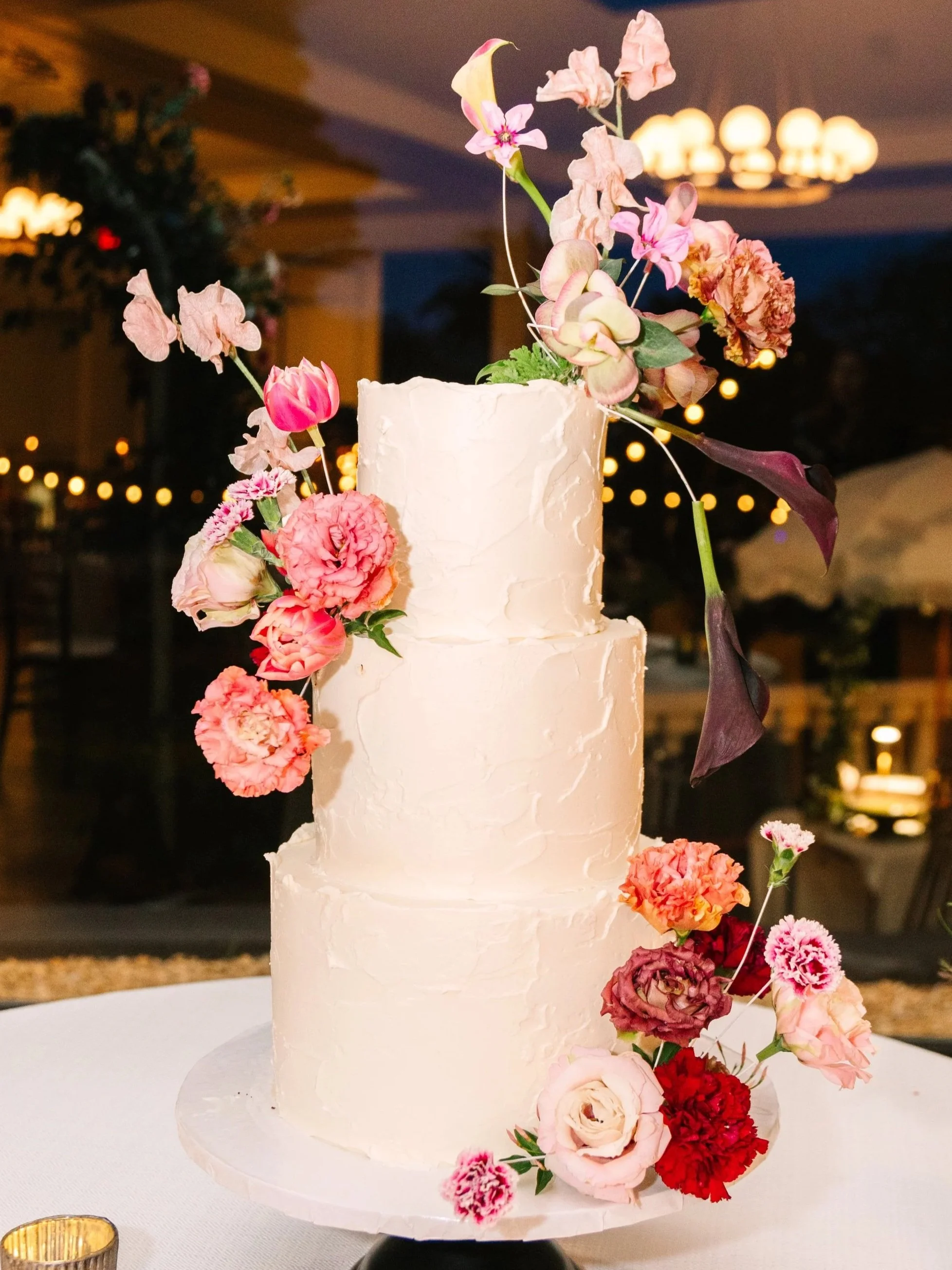 Three-tiered white wedding cake decorated with pink, red, and beige flowers, with some flowers cascading down the side, placed on a white tablecloth at an indoor event with warm lighting.