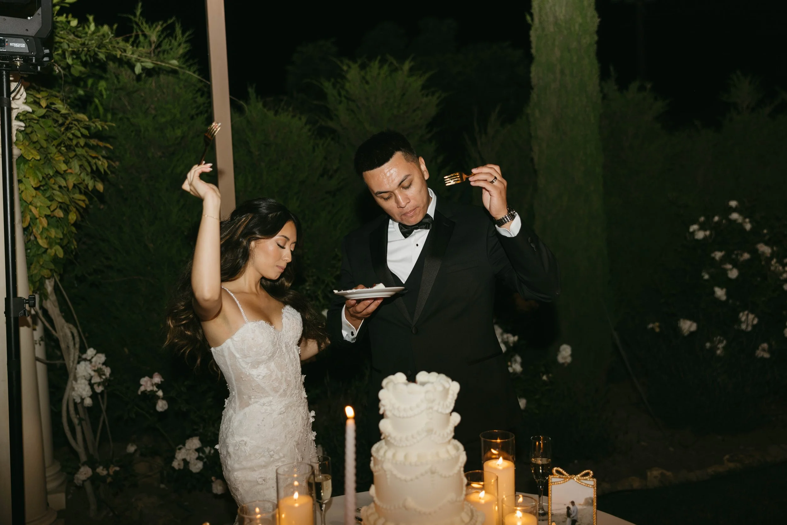 A bride and groom are standing at a wedding reception table with candles and a wedding cake, holding forks and appearing to be making a toast or listening to something. The bride is wearing a white lace wedding dress, and the groom is in a black tuxedo. They are outdoors at night surrounded by greenery and flowers.