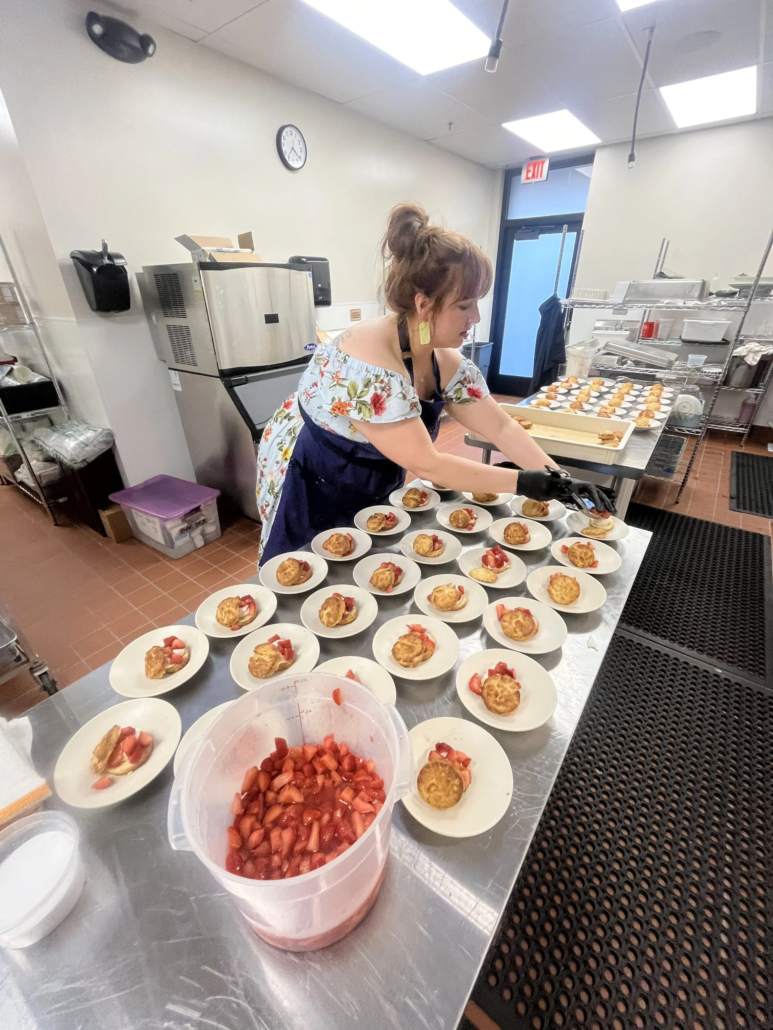 A woman preparing bite-sized desserts with strawberries and a pastry or puff pastry shell on a stainless steel table, with plates arranged with the same dessert and a bowl of chopped strawberries in a commercial kitchen.