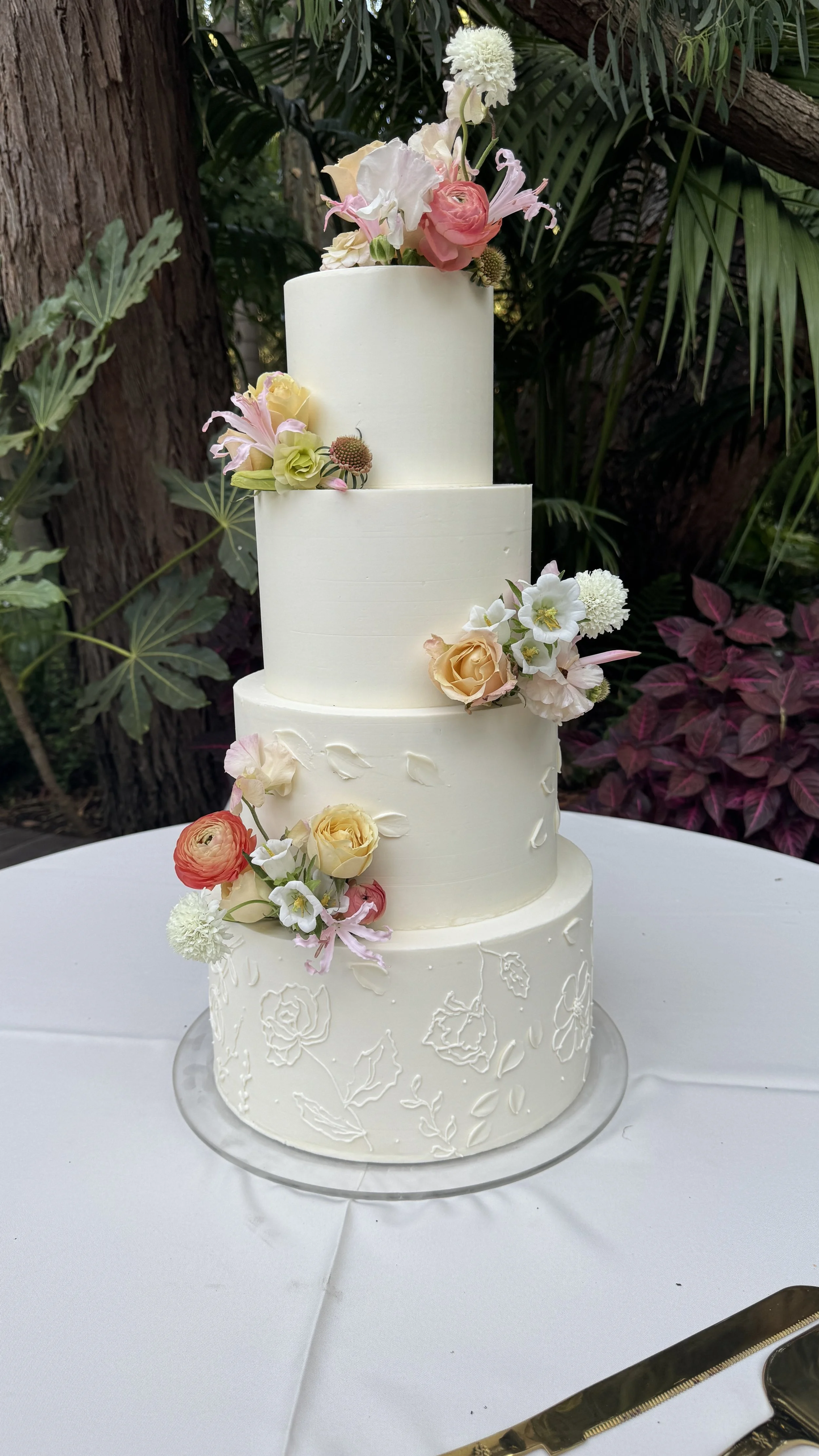A tall, three-tiered white wedding cake decorated with fresh pastel-colored roses, ranunculus, and other flowers, placed on a white tablecloth outdoors with green foliage in the background.