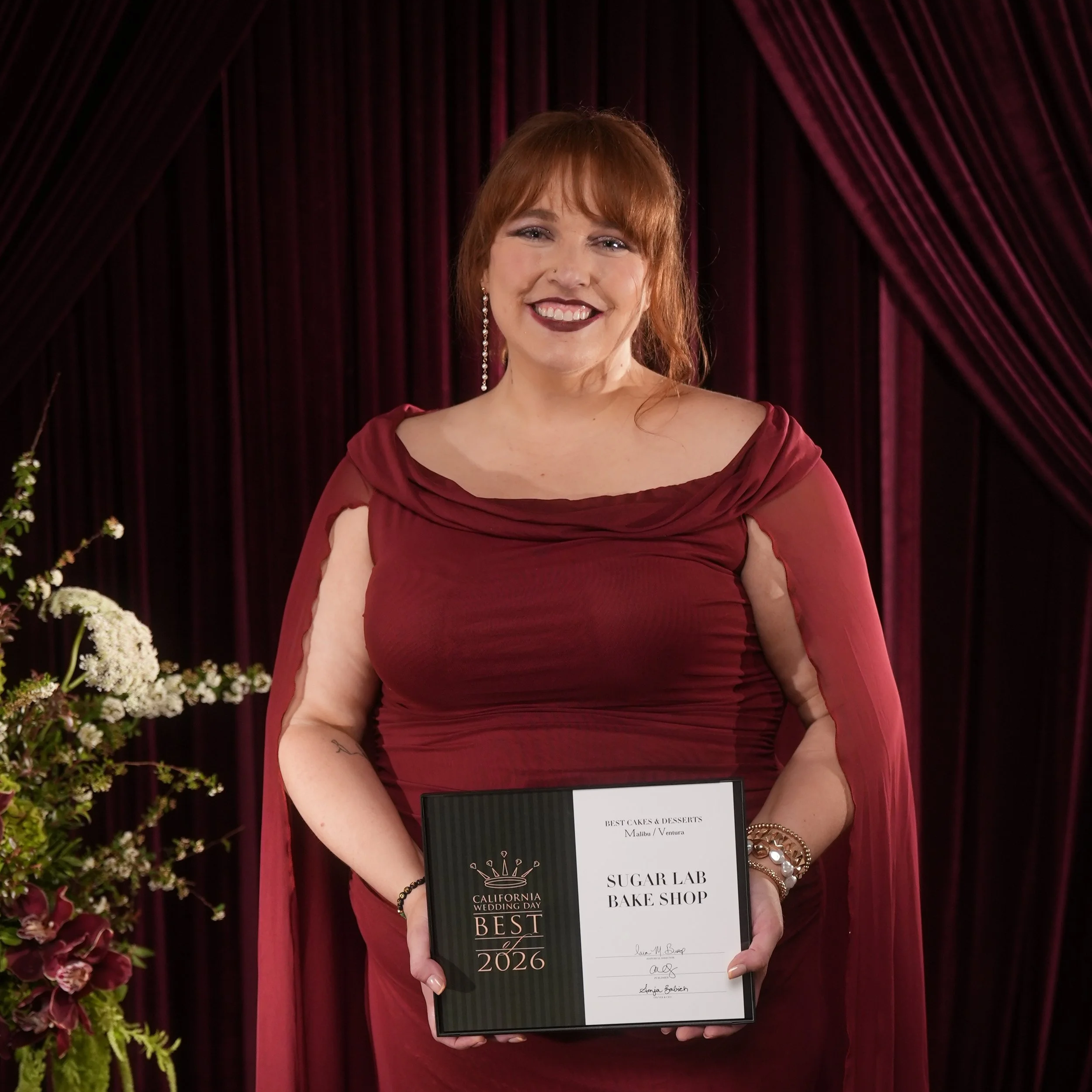 A woman dressed in a deep red gown holding an award plaque at a wedding event. The background has dark red curtains and floral arrangements.