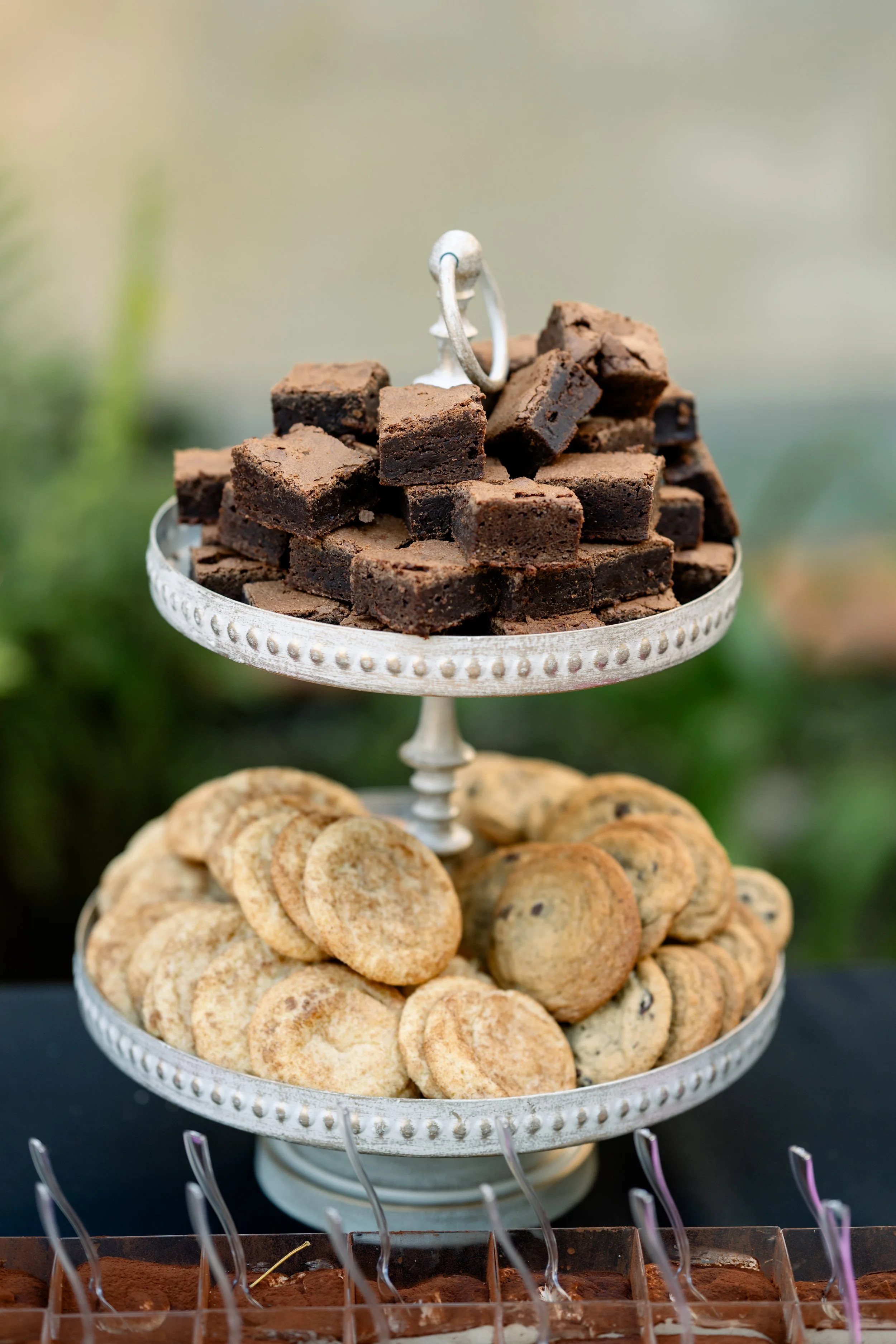 A three-tiered silver dessert stand filled with chocolate brownies on the top tier and assorted cookies on the bottom tier, with additional desserts visible beneath.