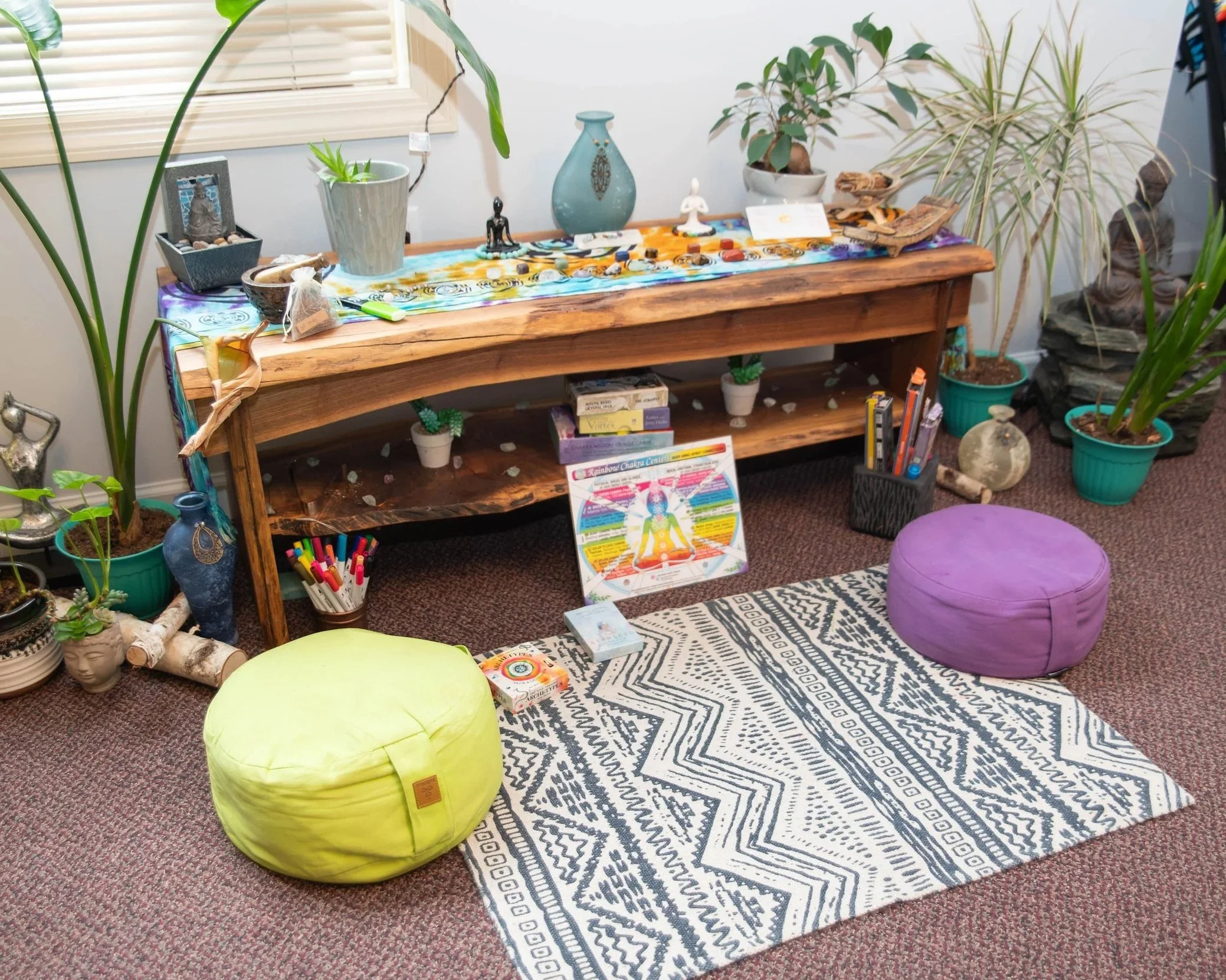 An indoor yoga or meditation space with a wooden table adorned with crystals, plants, and decorative items. On the floor, there is a black and white patterned rug with a yellow and a purple meditation cushion. The space includes indoor plants in various pots, books, markers, and a colorful chart related to energy or chakras. The room has a cozy, bohemian style.