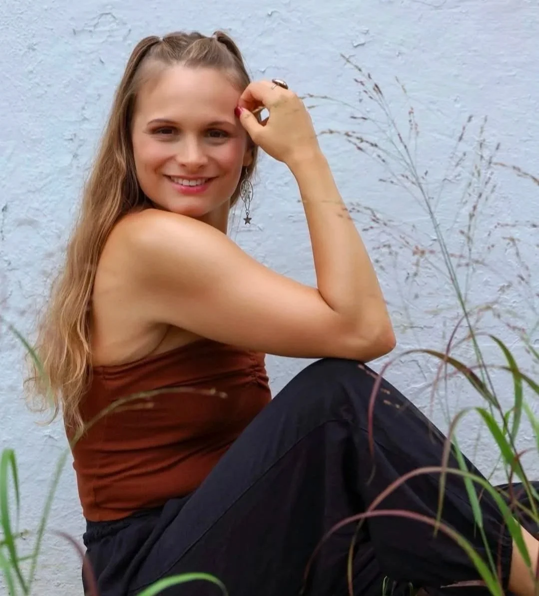 A woman with long, wavy blonde hair sitting outdoors against a textured white wall, smiling at the camera. She wears a strapless brown top and black pants, with star-shaped earrings.