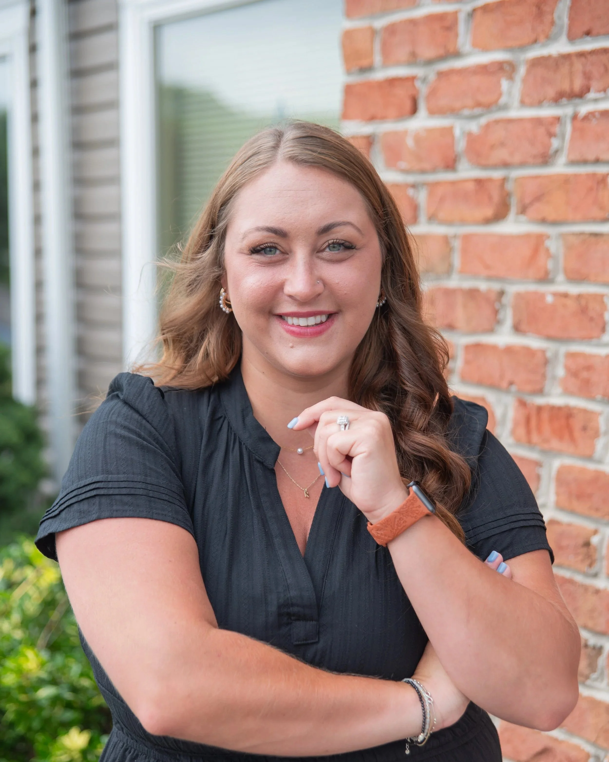 A woman with long wavy brown hair, wearing a black short-sleeved top, standing outdoors near a brick wall with greenery in the background, smiling and looking at the camera.