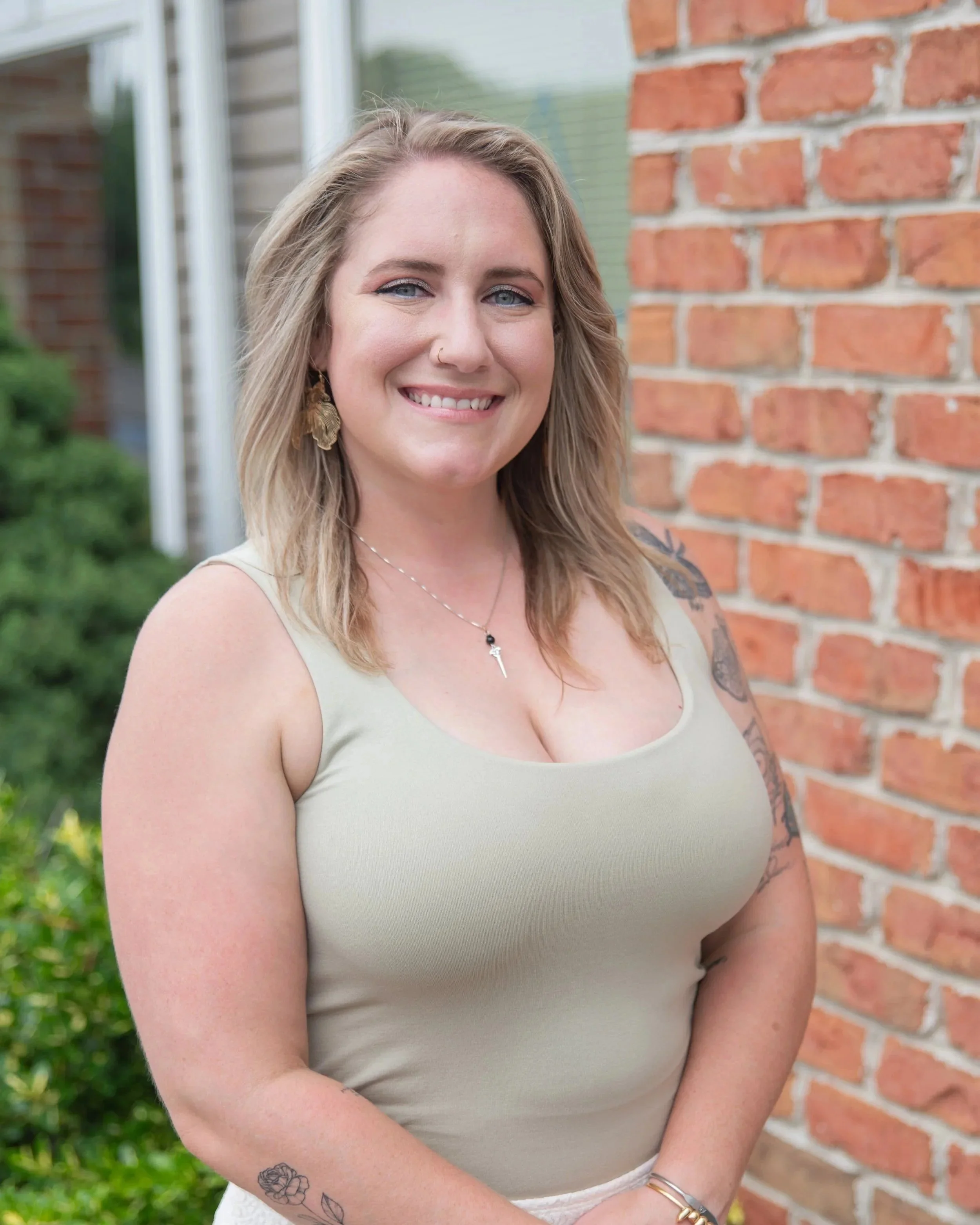 A woman with shoulder-length blonde hair and blue eyes smiling outdoors, standing in front of a brick wall and greenery.