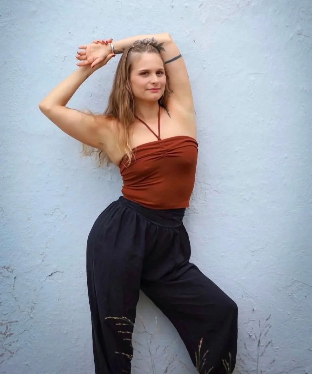 A woman with long, wavy hair and a tattoo on her upper arm stands against a light blue wall, posing with her arms raised and hands behind her head, wearing a strapless brown top and black pants.
