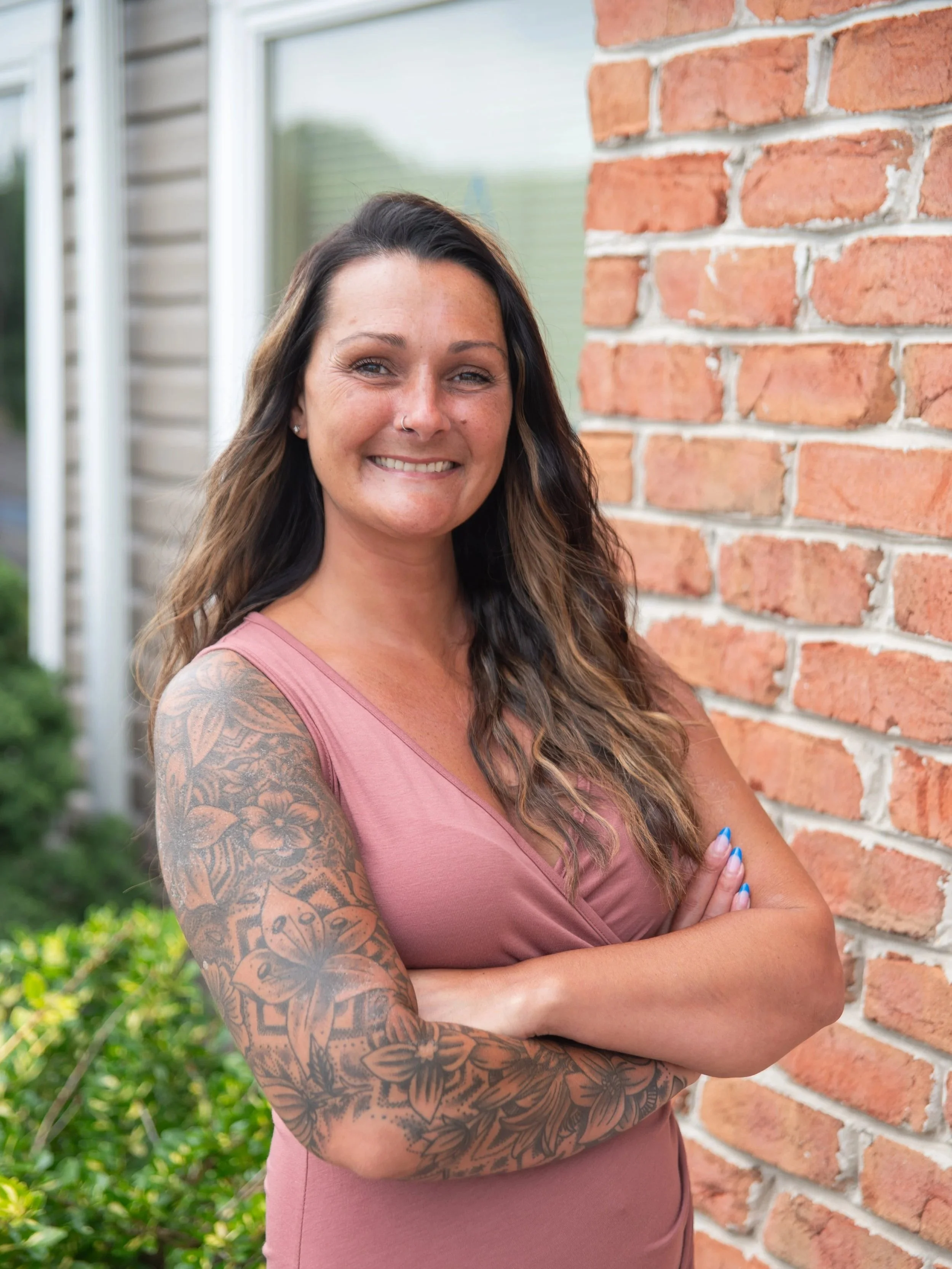 A woman with long wavy brown hair and tattoos on her left arm, smiling with crossed arms, standing outdoors near a brick wall and house.